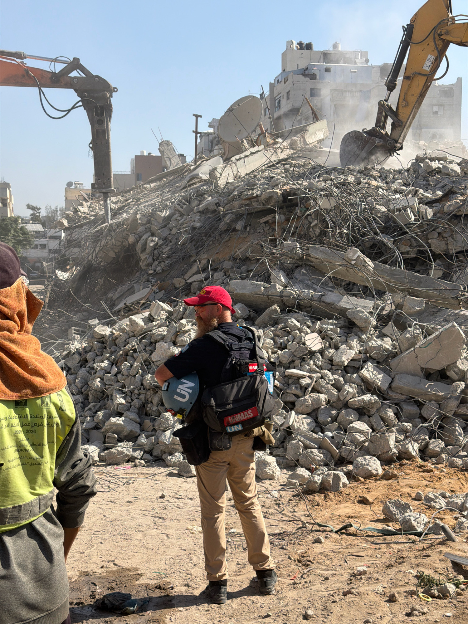 In Gaza City, Palestinian Territories, amidst a landscape of widespread destruction, a humanitarian worker in a bright yellow vest stands with their back to the camera, facing a colossal pile of rubble. The rubble, a chaotic amalgamation of concrete, twisted rebar, and debris, is the result of what appears to be a building demolition or destruction. Two excavators, one orange and one yellow, are visible in the background, actively engaged in the clearing or demolition process. The yellow excavator's bucket is lowered into the debris, raising a cloud of dust.
In the foreground, another individual, a male wearing a red baseball cap, a blue shirt, and tan pants, stands facing away from the camera, holding a light blue helmet with a UN logo. He is wearing a tactical vest with several pouches and bags attached. This individual appears to be part of a humanitarian or aid organization, likely involved in assessing the damage or assisting in the recovery efforts. The environment is dusty and dry, with a clear blue sky overhead. The backdrop features partially destroyed buildings, indicating the scale of the destruction in the area.