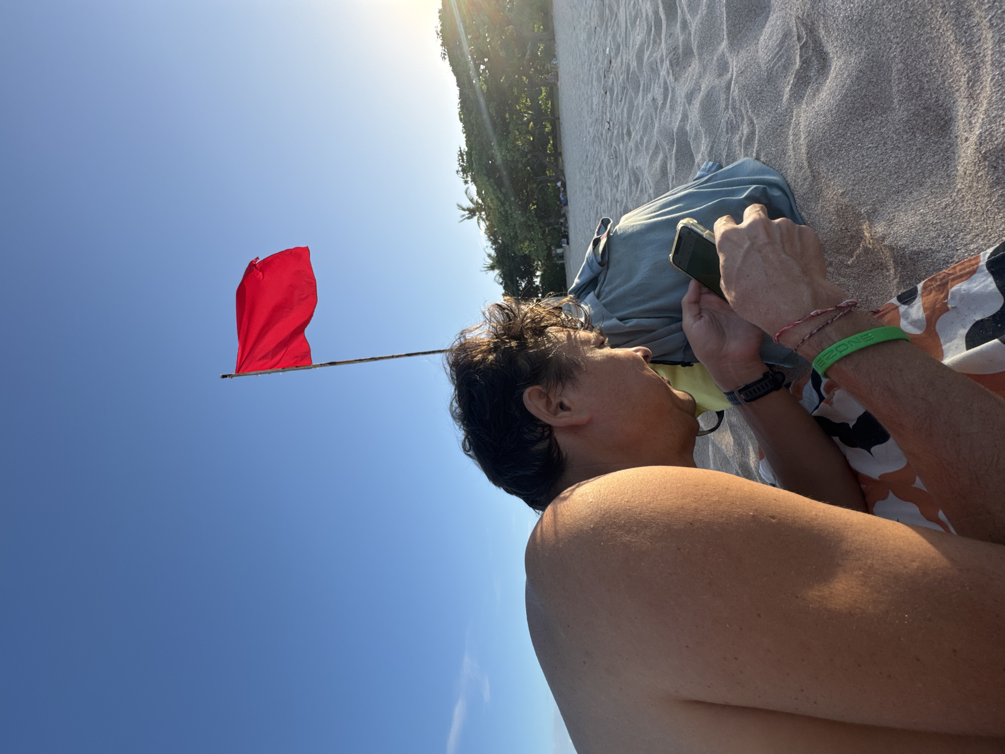 The image captures a man lying on a sandy beach in Kuta Selatan, Indonesia. He is seen from a low angle, positioned on his side, looking at his phone. He wears a green bracelet on his left wrist and a woven one on his right. A red flag is erected in the sand nearby. Behind him are trees and the sun is shining. The beach itself is light-colored sand.