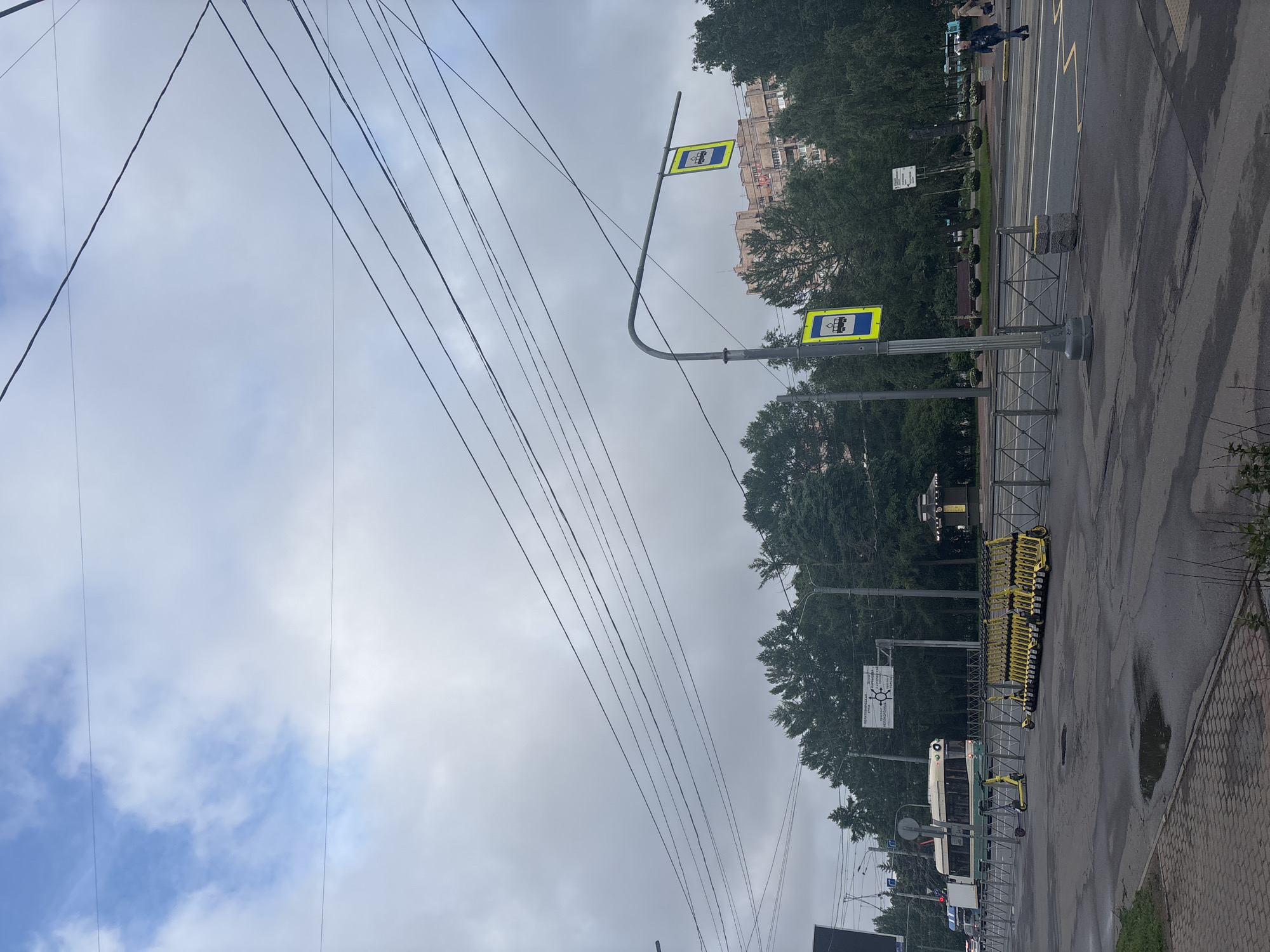 The image depicts a cloudy day in Saint Petersburg, Russia, on Politechnicheskaya Street. In the foreground, the asphalt street is wet, with puddles reflecting the sky. A row of yellow electric scooters is parked along a metal fence. Further down the street, a tram is visible. The background features a dense line of green trees, behind which a multi-story residential building stands. Two bus stop signs are prominently displayed on poles. In the distance, a person is walking across the street. Overhead, numerous power lines crisscross the sky, obscuring some of the clouds.