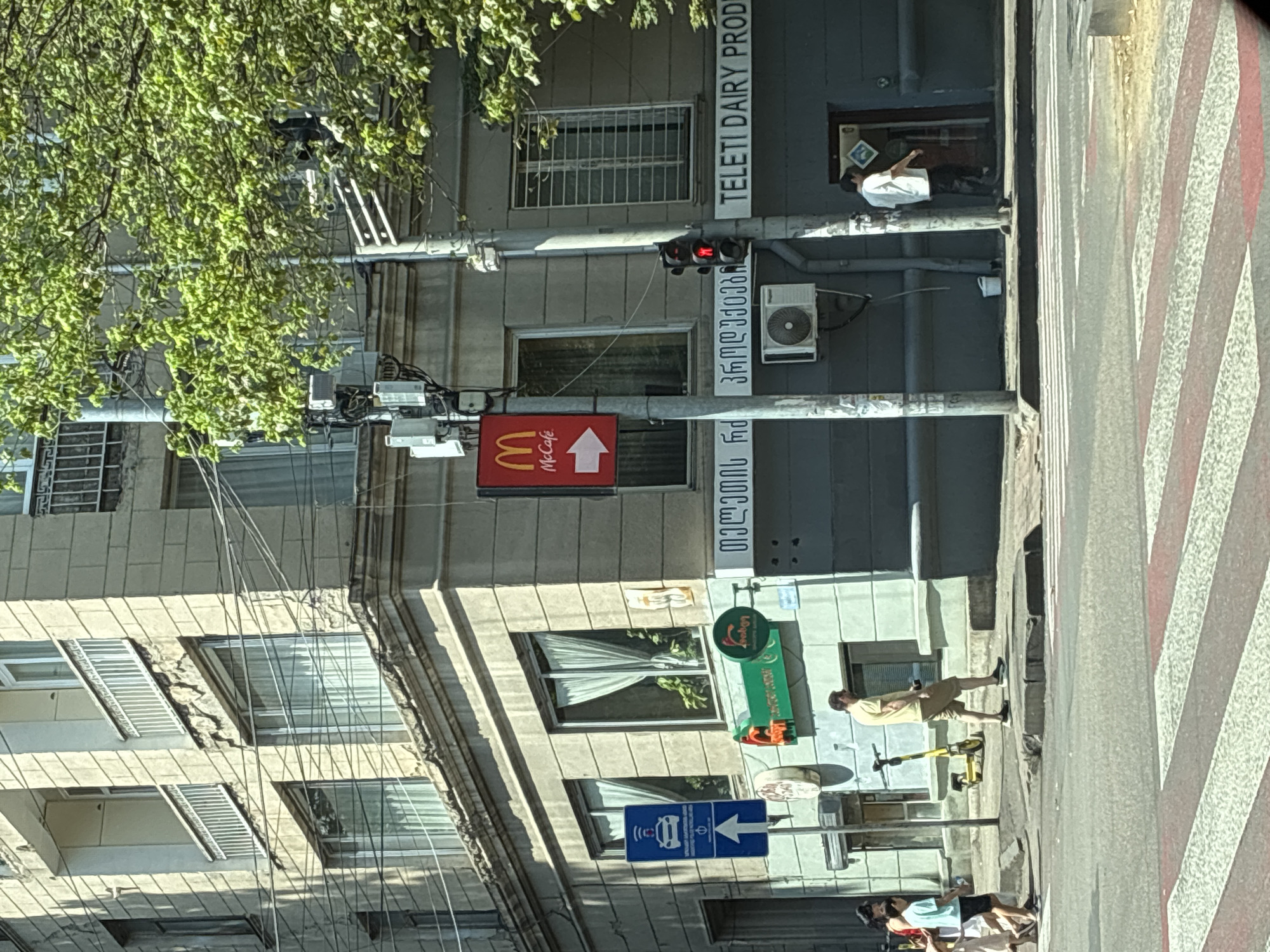 The image captures a street scene in Tbilisi, Georgia, specifically at an intersection. A multi-story building, weathered and with visible signs of age, forms the backdrop. The building features balconies with white railings and numerous windows, some covered with sheer curtains. On the exterior of the building, several signs are visible. A prominent red and white McCafe sign with an arrow points to the right, indicating the direction of a McDonald's establishment. Below this, there's signage in what appears to be Georgian script. To the left, a blue traffic sign displays a car icon with a wifi symbol above it and an upward-pointing arrow. Another green sign with text is also present.
The street is paved, and a pedestrian crossing with red and white stripes is clearly marked. Several individuals are present. On the sidewalk to the left, a group of people, including children, are walking. A man in a light yellow t-shirt and beige shorts walks away from the camera. A yellow electric scooter is parked on the sidewalk. Further down the street, two men are visible near an entrance. One man, wearing a white t-shirt and dark pants, stands by a doorway. Another man in a white shirt stands inside what appears to be a shop. An air conditioning unit is mounted on the building wall. Traffic lights are visible, one with a red light illuminated. The environment is bright and appears to be daytime, with green trees overhanging part of the scene. The overall impression is that of a busy urban intersection.