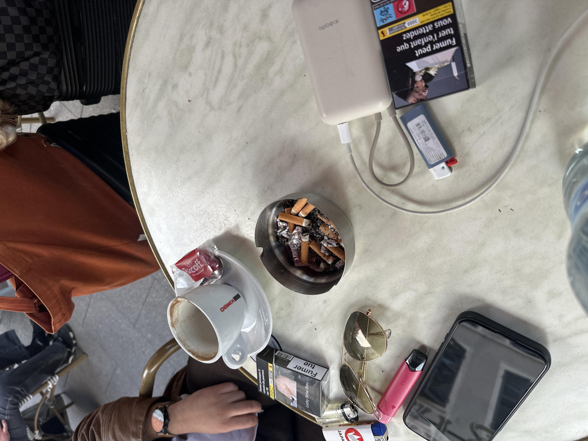 The image depicts an overhead shot of a round, faux-marble table at an outdoor café, possibly in Strasbourg, France, given the French language on the cigarette packaging. The table is cluttered with various items, suggesting a person has been sitting there for some time.
Centered is an ashtray filled with numerous cigarette butts. To its left is a white coffee cup with the brand name KIMBO visible and a Biscoff cookie wrapper on the saucer.
To the right of the ashtray are sunglasses, a pink vaping device, and a smartphone. Adjacent to these items are a pack of Lucky Strike Bleu cigarettes and two lighters.
At the top of the frame is a Xiaomi power bank connected with a charging cable. A pack of cigarettes with a warning label in French is also present. A bottle of water is barely visible on the upper right.
The edge of a suitcase is seen on the left, indicating someone's luggage. A person's hand with a wristwatch is visible in the bottom left, suggesting the person is seated at the table. The person is wearing a brown jacket. An orange article of clothing and a portion of a chair are also visible near the edge of the table. The surface of the table is aged, with a slightly cracked surface.