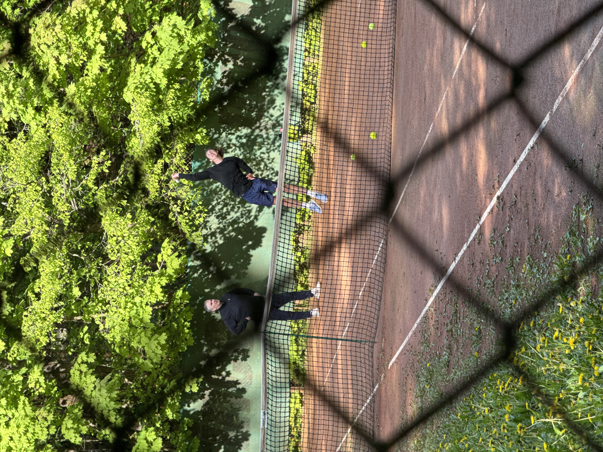 The image depicts a tennis court viewed through a chain-link fence. Two men are on the court. One man is wearing a black long-sleeved shirt and dark blue shorts and white shoes. The second man is wearing all black clothing and white shoes. The tennis court is brown with white lines and has a green net. There are several tennis balls on the court. A tree with green leaves is visible on the left side of the image. The location is Unknown, Russia.