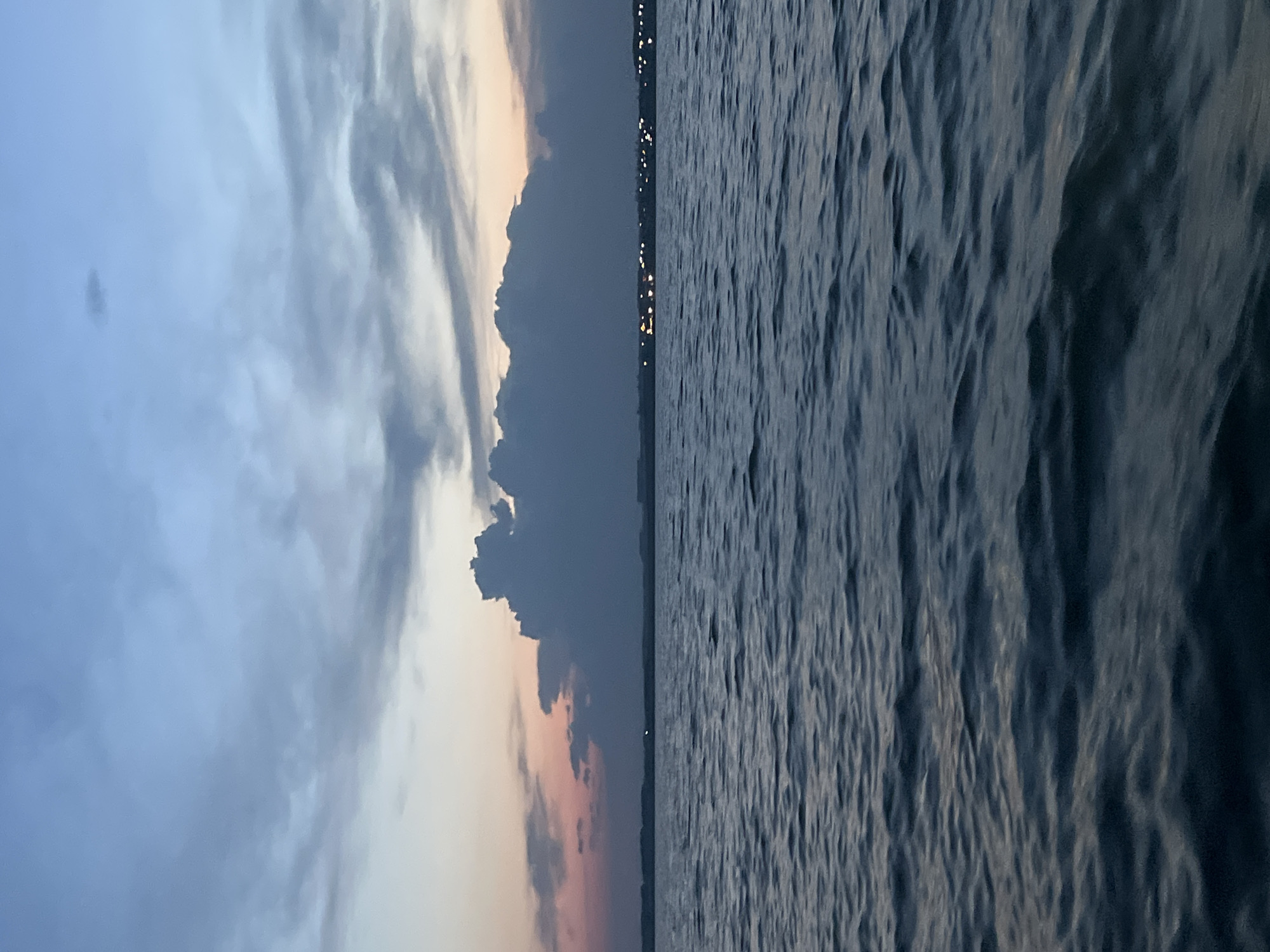 The image depicts a serene body of water under a twilight sky, possibly at dusk or dawn. The water's surface is rippled, suggesting a gentle breeze. In the distance, a faint line of lights indicates a settlement on the horizon. A large, dark cloud formation dominates the middle ground of the sky, with lighter clouds streaked across the upper portion. The colors in the sky range from pale blues and grays to soft pinks and oranges near the horizon, indicative of the fading or emerging sunlight. No people or specific objects are discernible in the scene. The location is identified as Novopichugovo, Russia, suggesting this is a view of a lake or bay in that area.