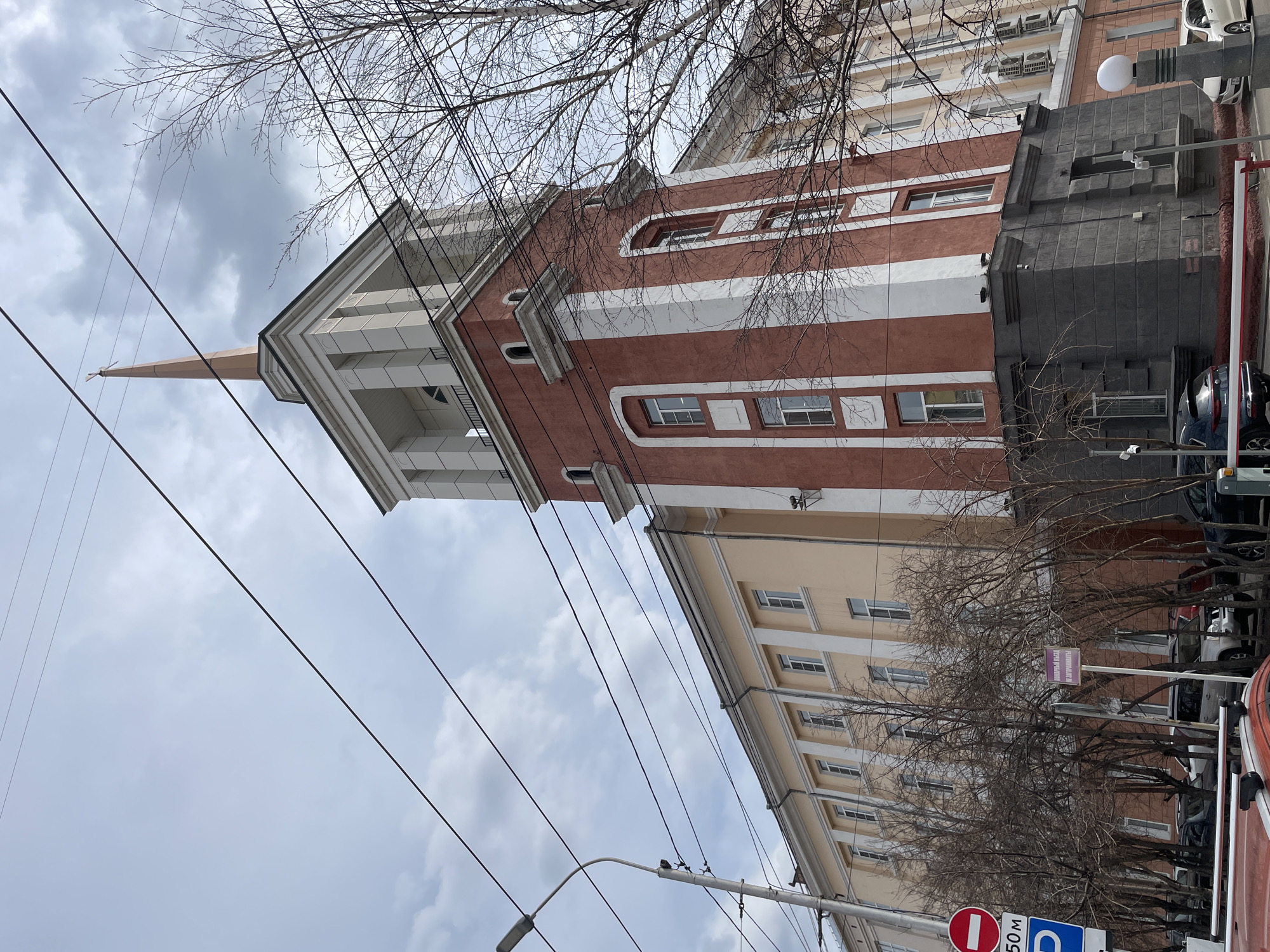 The image shows a building in Krasnoyarsk, Russia, notable for its striped red and white tower topped with a spire. The building is adjacent to another multi-story building of light yellow color. Bare tree branches and utility wires fill the upper portion of the frame, obscuring the sky with overcast conditions. At the street level, there are trees, parked vehicles, and street signs, including a parking sign.