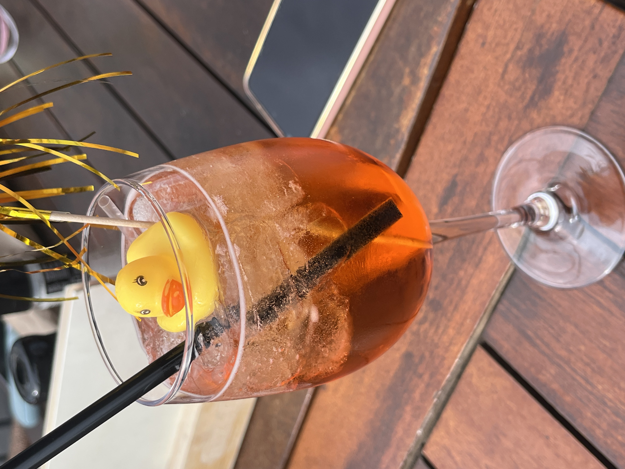 A high-angle, tilted shot depicts an Aperol spritz in a stemmed glass, garnished with a yellow rubber duck and gold tinsel. The drink contains ice and is served with a black straw. The glass rests on a brown wooden surface. The top edge of a mobile phone is visible in the background. The image suggests an outdoor setting, possibly a cafe or bar in Cochem, Germany.