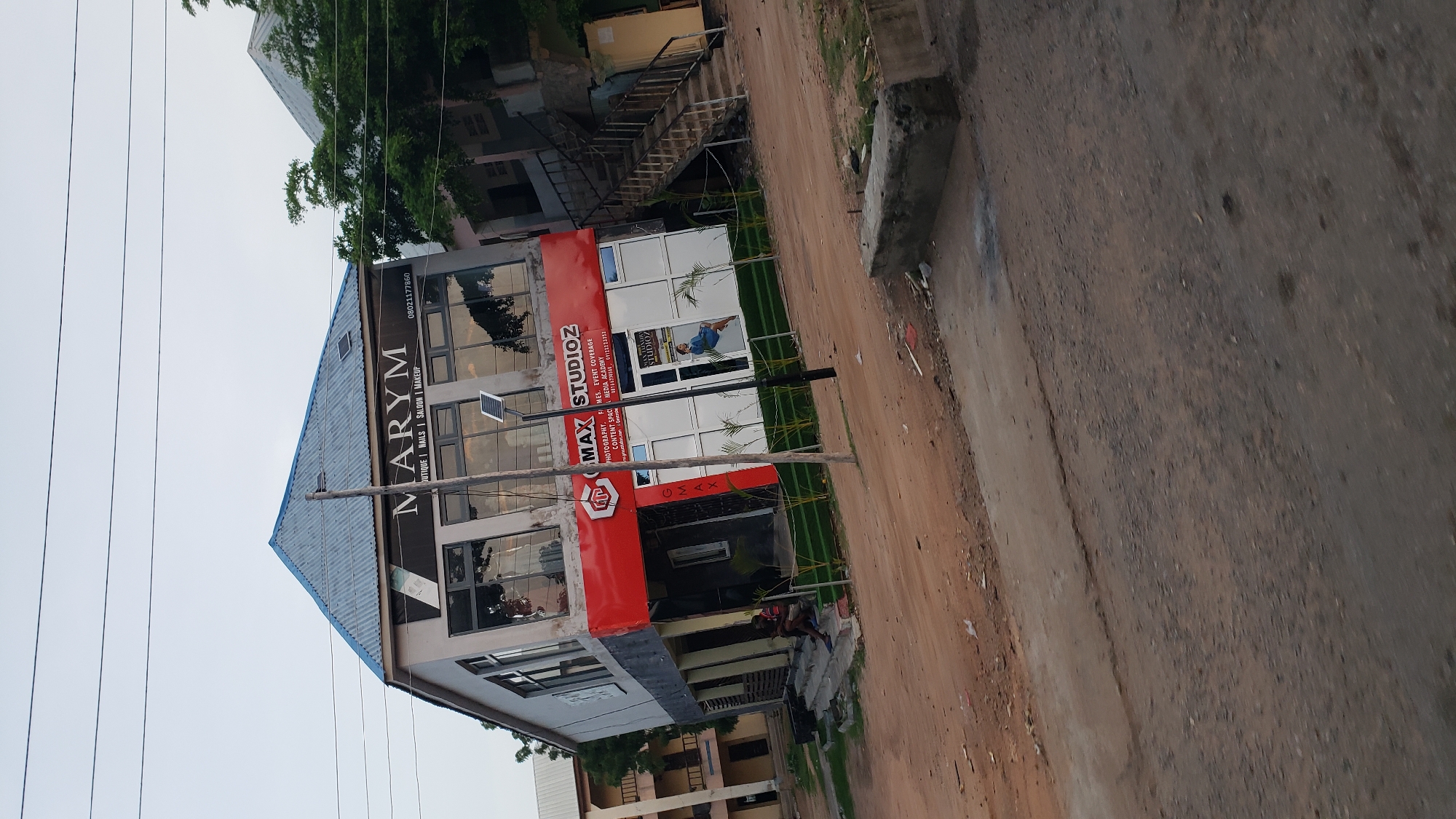 In Jalingo, Nigeria, a commercial building stands on a dirt road. The building features a blue corrugated metal roof and a grey facade. The lower level has a large red awning with white lettering that reads "G MAX STUDIOZ". Below this, a glass storefront displays advertisements, one featuring a woman in a blue dress. Several people are visible sitting on the steps leading up to the entrance. A solar-powered street lamp is positioned in front of the building, and a few potted plants are lined up along the storefront. In the background, other buildings and trees can be seen. The ground in front of the building is unpaved and covered in dirt and gravel. Power lines are visible overhead.