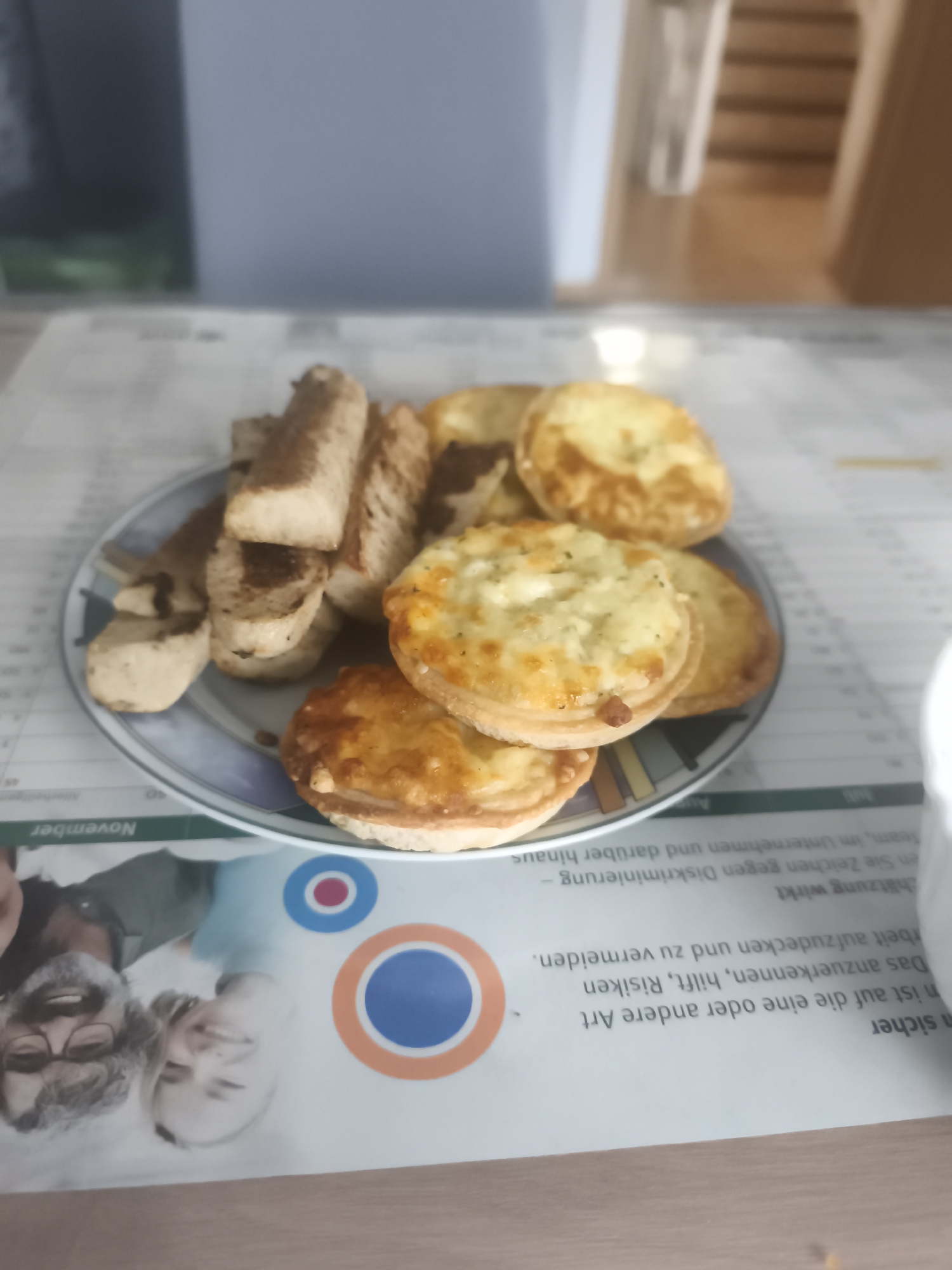 The image shows a plate of food, likely a snack or light meal, placed on a table. The food consists of several pieces of toasted bread or bruschetta, and small, round pastries filled with a creamy mixture, possibly cheese or a savory filling. The plate has a decorative rim with blue, purple, and white patterns.
The setting appears to be indoors, possibly a home or office, as indicated by the blurry background which includes furniture and a wall. The table is covered with a sheet of paper that has printed text and images. The text, partially visible and oriented at an angle, is in German and discusses topics like "Diskriminierung" (discrimination), "Risiken" (risks), and "Unternehmen" (companies), suggesting it might be a document related to business or human resources. A part of the word "November" is also visible on the paper. There are also circular graphic elements with red, blue, and orange colors, and a faded image of two people, a man and a woman, with smiling expressions.
The lighting suggests it could be daytime, but no specific time can be determined. The overall mood is casual and appears to be a moment of pause or a break. The focus is primarily on the food, which is presented in a way that suggests it's ready to be eaten.