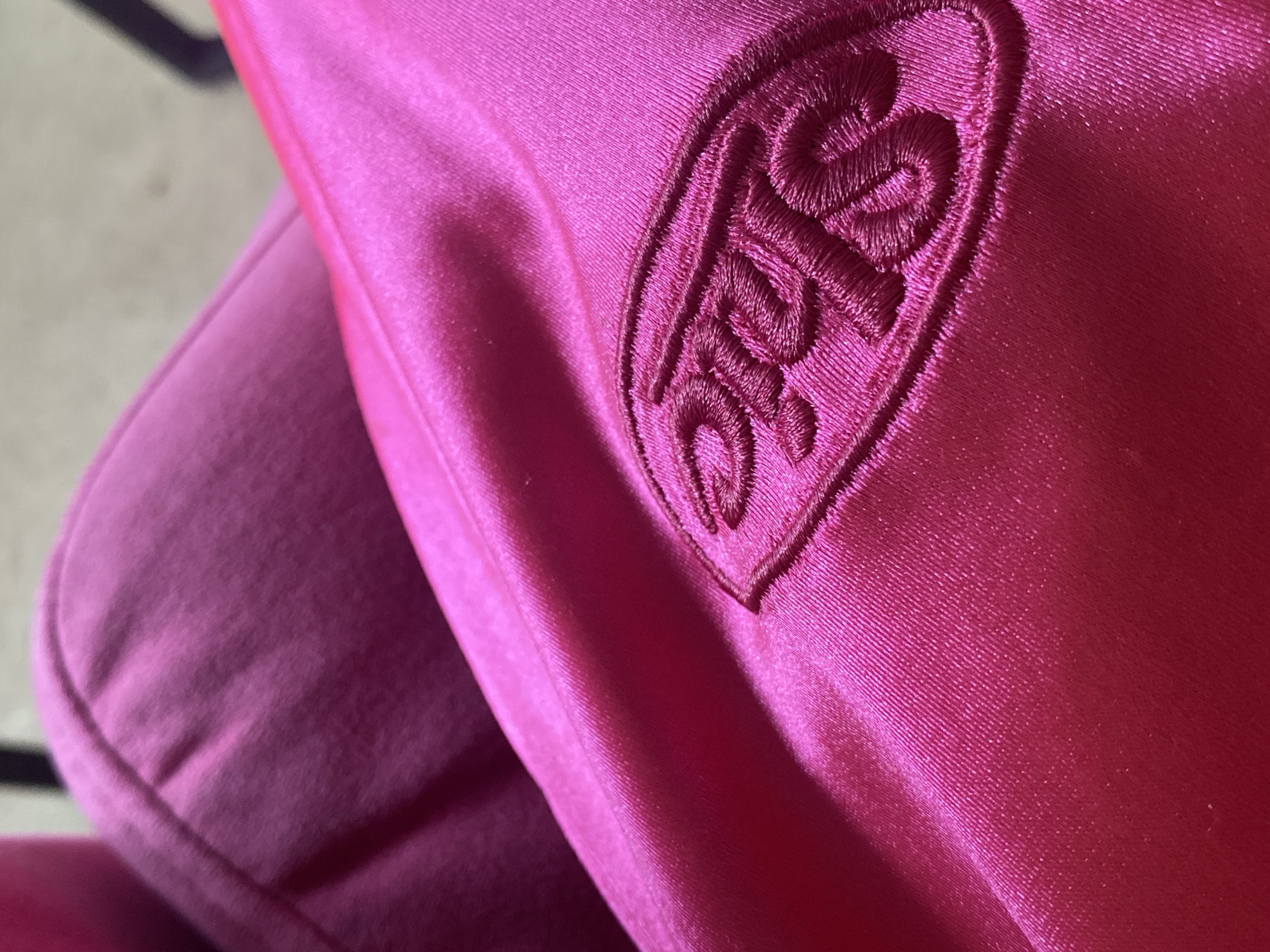 A close-up photograph shows a pink garment with an embroidered heart-shaped logo on its upper-right side. The logo features stylized lettering. The garment appears to be made of a smooth, possibly satin or similar fabric. Another part of the garment, made of a soft material, is visible in the background on the left. No information about the location, including Votkinsk, Russia, can be deduced from the image.