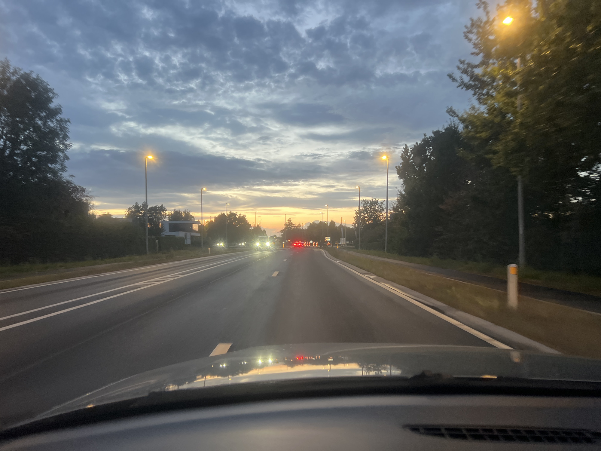 The image is a first-person perspective shot from inside a car, driving on a multi-lane road at dusk. The sky is filled with scattered clouds, illuminated by the setting sun which casts a warm glow on the horizon. Streetlights along the road are lit, as are the headlights of oncoming vehicles. Trees line both sides of the road, with some modern buildings visible in the background on the right. The road has clear white lane markings.