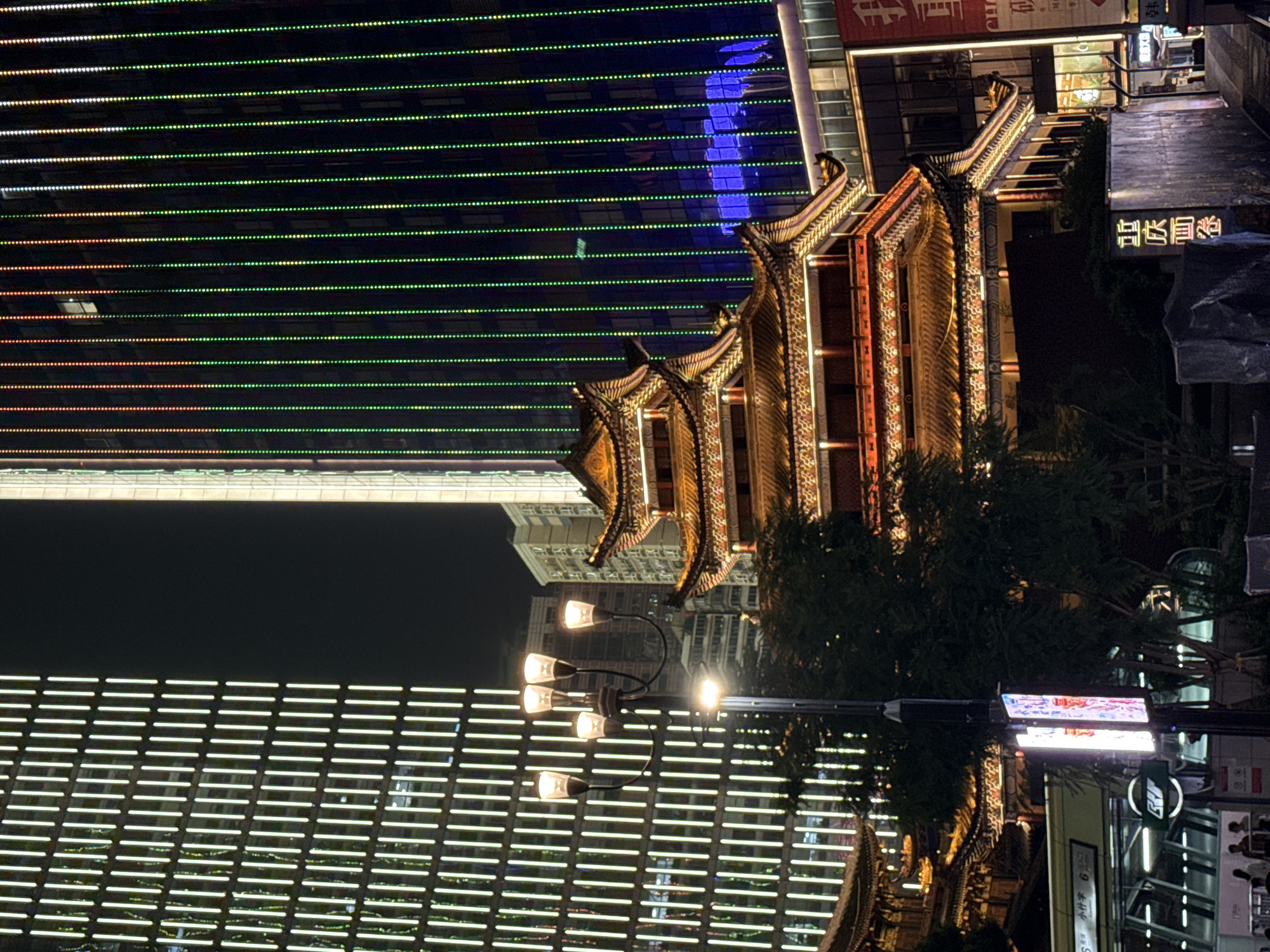 The image depicts a nighttime view of a cityscape in Yuzhong District, China, characterized by a juxtaposition of modern architecture and traditional Chinese elements. Dominating the scene are two tall, modern skyscrapers. The building on the left is illuminated with vertical white lights, creating a grid-like pattern across its facade. The skyscraper on the right is adorned with vertical rows of colorful LED lights that transition from red at the top to green lower down.
In the foreground and mid-ground, a traditional multi-tiered Chinese building, possibly a temple or cultural center, is brightly lit with warm orange and yellow lights. Its distinctive curved rooflines and ornate details are clearly visible. A street lamp with four illuminated fixtures stands between the traditional building and the modern skyscrapers. In front of the traditional building, a lush dark green tree partially obscures the view. Further in the foreground, there are hints of people walking and what appears to be an entrance to a subway station or public transport hub with signage. The overall atmosphere is one of vibrant urban nightlife, where ancient architectural styles are preserved amidst contemporary development.