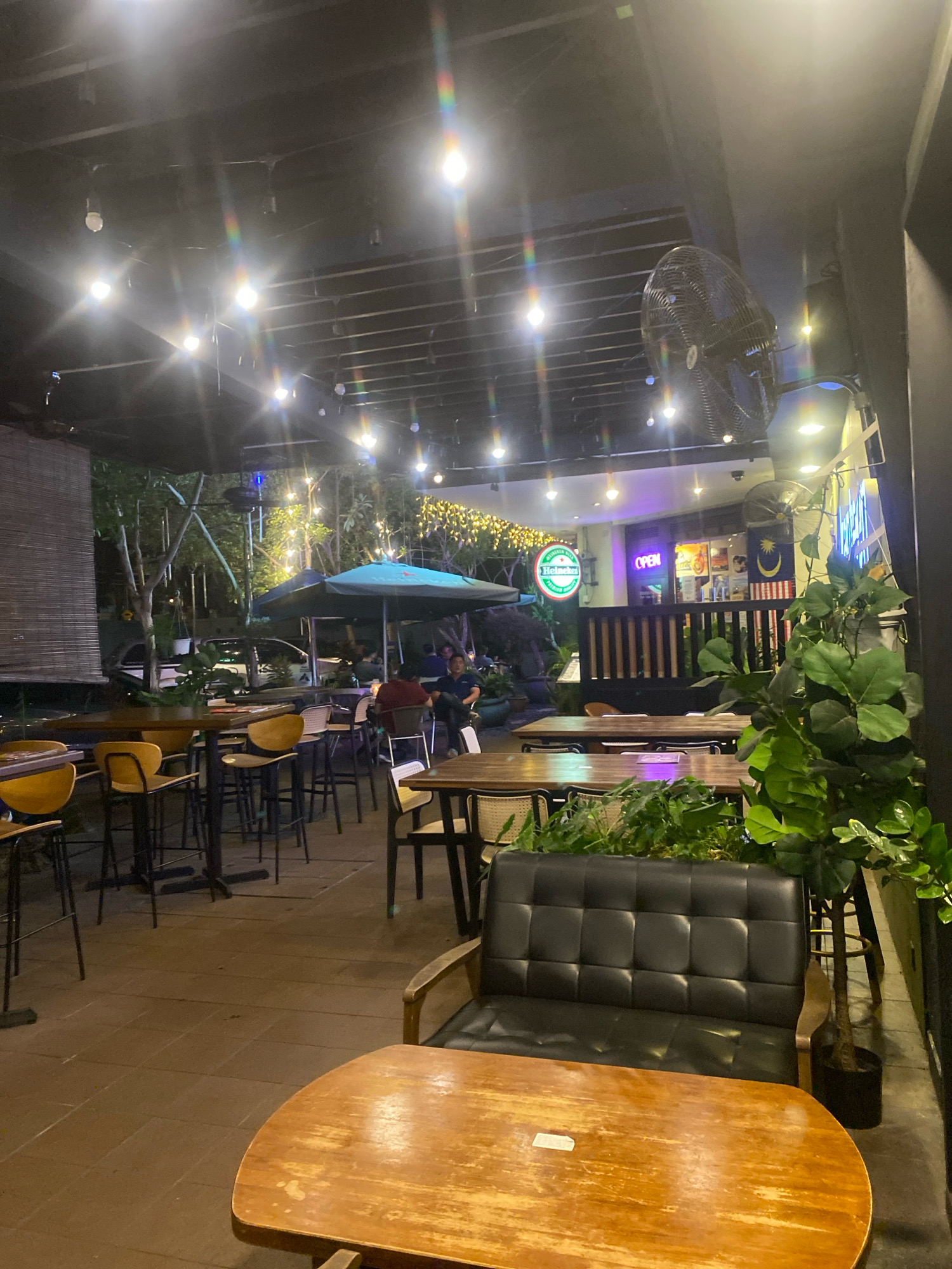 The image shows an outdoor seating area of a restaurant or cafe, likely in Kuala Lumpur, Malaysia, given the presence of the Malaysian flag. The scene is dimly lit by string lights and spotlights, creating a warm ambiance. In the foreground, a wooden table is visible, with a black leather sofa to its right. Further back, there are rows of tables and chairs, some of which are occupied by patrons. A large green umbrella provides shade over one of the seating areas. In the background, a store with an "OPEN" sign is visible, along with a large industrial fan mounted on the ceiling. The overall setting suggests a casual dining or social gathering space.