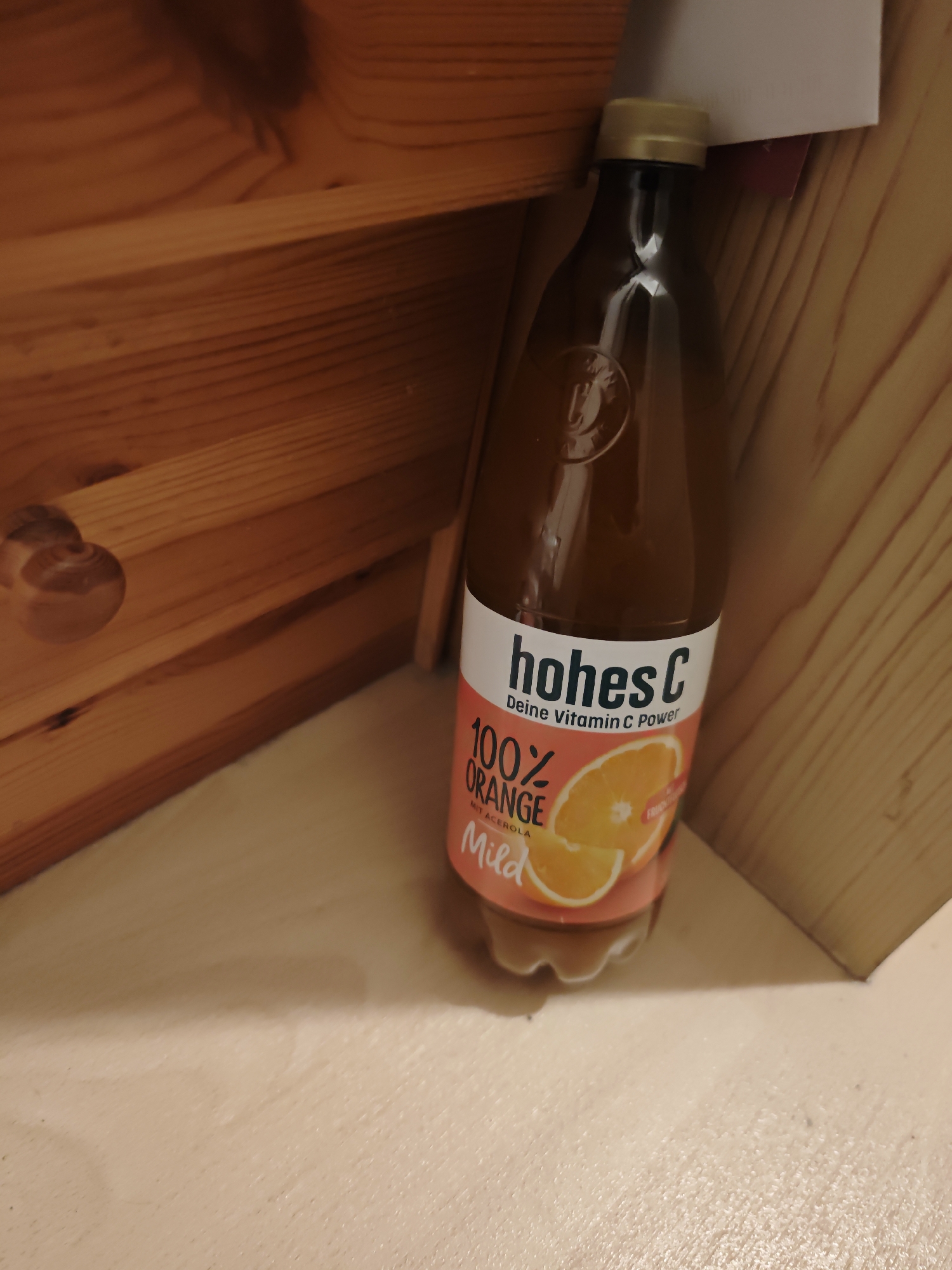 A close-up, low-angle shot features a brown glass bottle of "hohes C" orange juice. The bottle has a gold cap and a label that reads "hohes C Deine Vitamin C Power 100% ORANGE MIT ACEROLA Mild." The bottle is positioned on a light-colored, textured surface, possibly a shelf or tabletop, in front of a wooden structure with horizontal planks. The wooden structure appears to be a shelf or cabinet with a decorative carved wooden circle on the left side. The lighting suggests an indoor environment, and the scene is framed to emphasize the product. No people or actions are visible. The location in Zweibrücken, Germany, is not determinable from the image itself; this information is external to the visual content.
