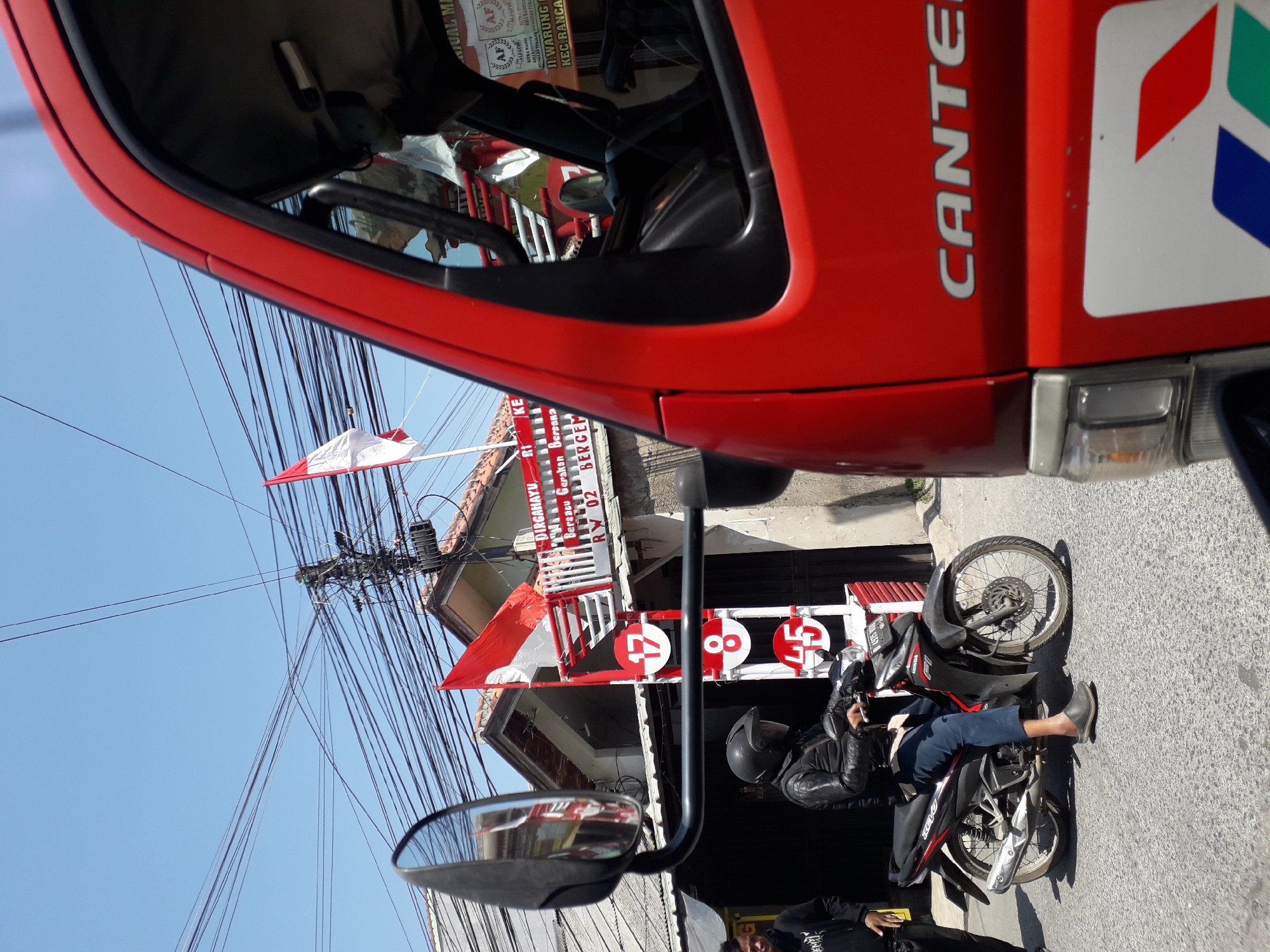 The image, taken in Linggar, Indonesia, depicts a street scene with a red truck in the foreground. A motorcycle with a rider wearing a black helmet and leather jacket is stopped on the right side of the frame. The rider is wearing blue pants and grey slip-on shoes. To the left of the motorcycle, a person wearing a black hoodie and black pants stands on the sidewalk. Behind the motorcycle, a building with red and white striped railings is visible. Two Indonesian flags, one red and white and another smaller red flag, are displayed on the building. A sign on the building displays the numbers "17", "8", and "45" within red circles. The sky is clear and blue, and a complex network of electrical wires is strung across the scene. The truck in the foreground has the word "CANTER" visible on its side.