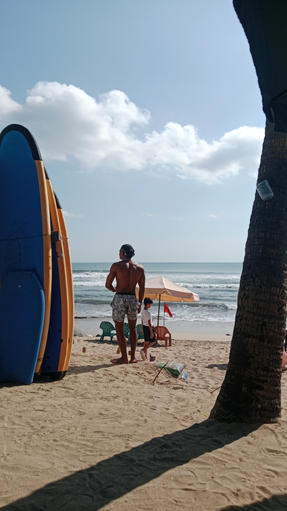 A medium shot captures a sun-drenched beach scene with a focus on a shirtless man facing away from the camera, looking out at the ocean. He is wearing patterned shorts and stands in the foreground, with two blue and yellow surfboards leaning against him on the left. A young boy in a white t-shirt and dark shorts walks past him towards the right. In the background, a beige umbrella provides shade over two empty chairs. The ocean stretches out to the horizon under a bright blue sky dotted with scattered white clouds. A palm tree trunk dominates the right side of the frame, casting a long shadow across the sandy beach. The overall environment suggests a tropical or coastal vacation setting.