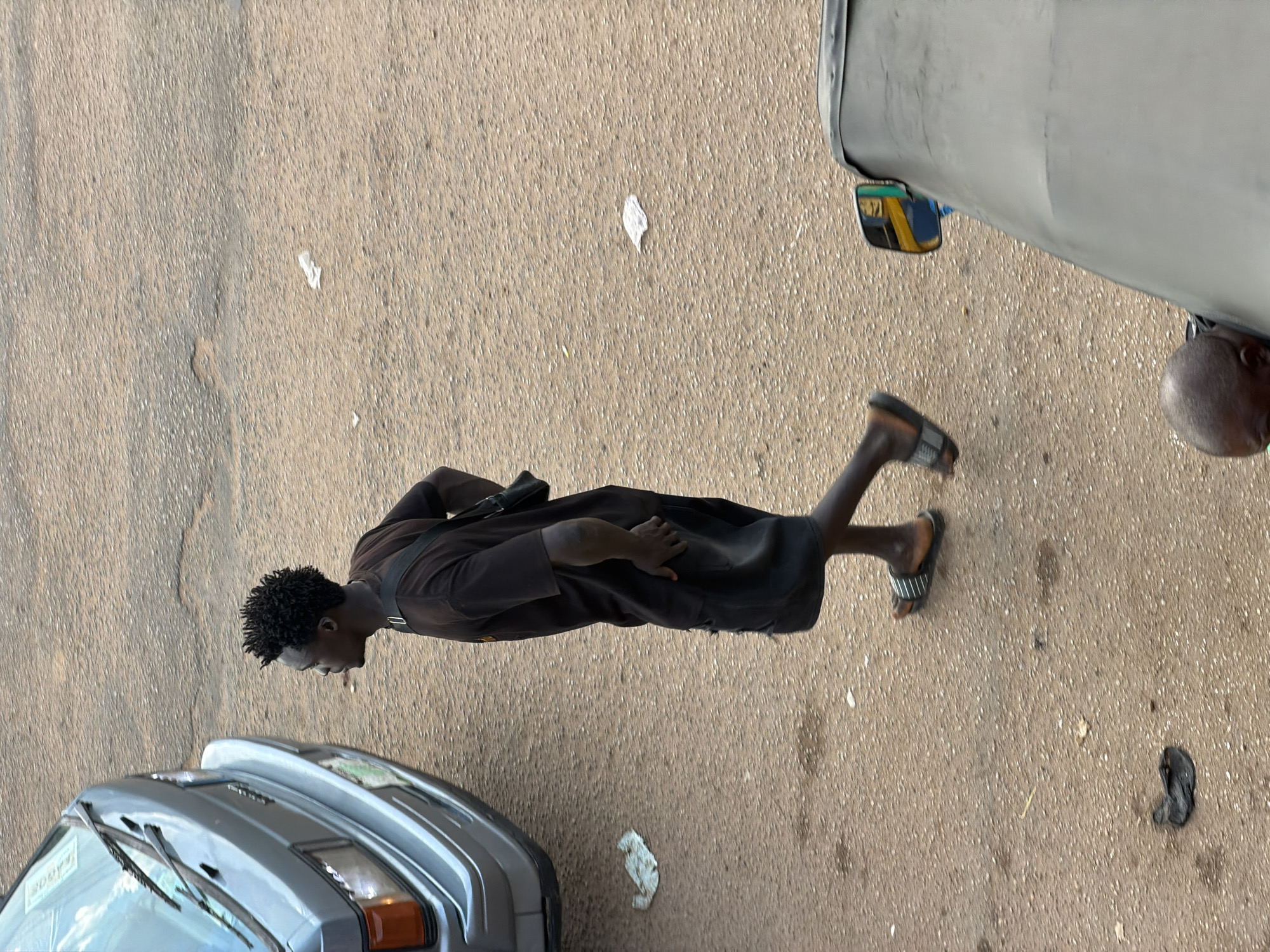 In Bukuru, Nigeria, a man in a dark t-shirt and shorts walks along a paved road. He has short, curly black hair and is wearing black sandals. A dark bag is slung across his chest. To the left of the man is the front of a grey vehicle, and to the right, part of a light-colored vehicle is visible, including a side mirror. The ground is a mix of asphalt and gravel, with some scattered debris. The scene appears to be outdoors during the daytime.