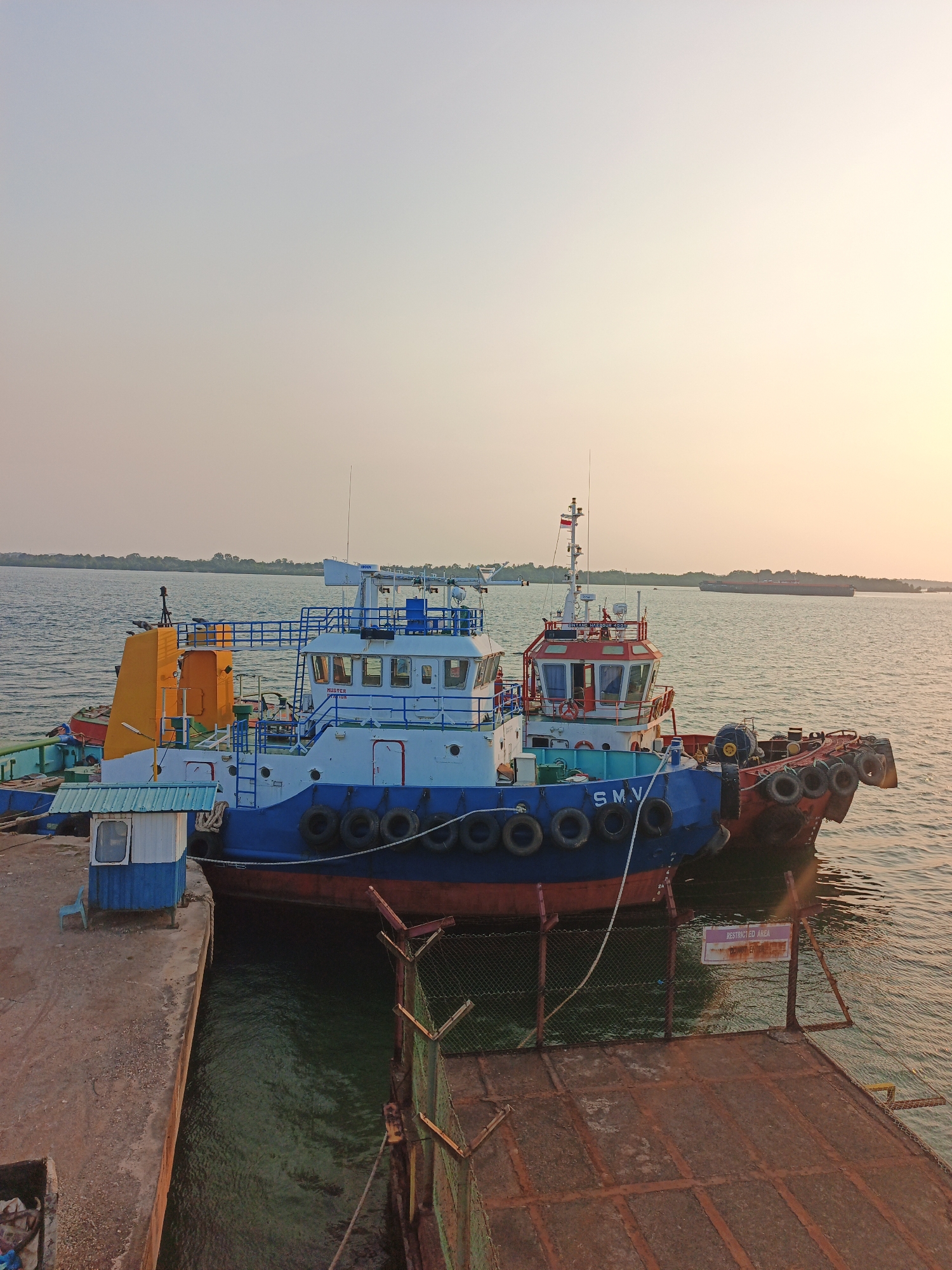 Two tugboats are docked side-by-side at a concrete pier in Batam, Indonesia. The tugboat on the left has a blue and white superstructure with a yellow crane at the rear. The tugboat on the right has a red and white superstructure. Both boats have a series of black tires mounted along their sides for fendering. A small blue and white booth with a single chair is positioned on the pier next to the left tugboat. A chain-link fence with a "RESTRICTED AREA DO NOT ENTER" sign is visible along the edge of the pier. In the background, a large barge is visible on the water, and a treed shoreline stretches across the horizon. The sky is clear and hazy, with the warm glow of sunset illuminating the scene. There are no people visible in the image.