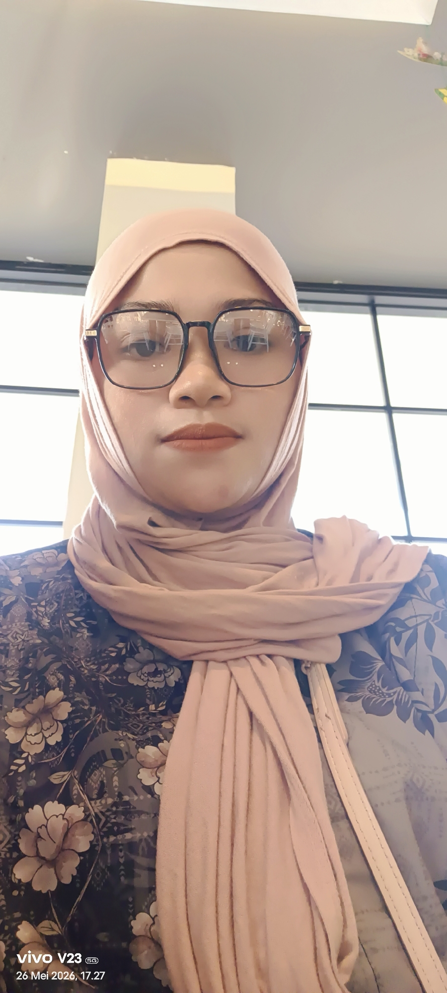 A medium-angle close-up photograph shows a woman wearing a pink hijab and black-framed glasses. She is facing the camera. The woman is wearing a dark floral print blouse. A light-colored strap runs diagonally across her torso. The background includes a window with vertical panes and part of the interior ceiling. A watermark on the bottom left corner of the photo reads "vivo V23 5G, 26 Mei 2026, 17.27." The location in the city of Lhokseumawe, Indonesia is not discernible from the image.