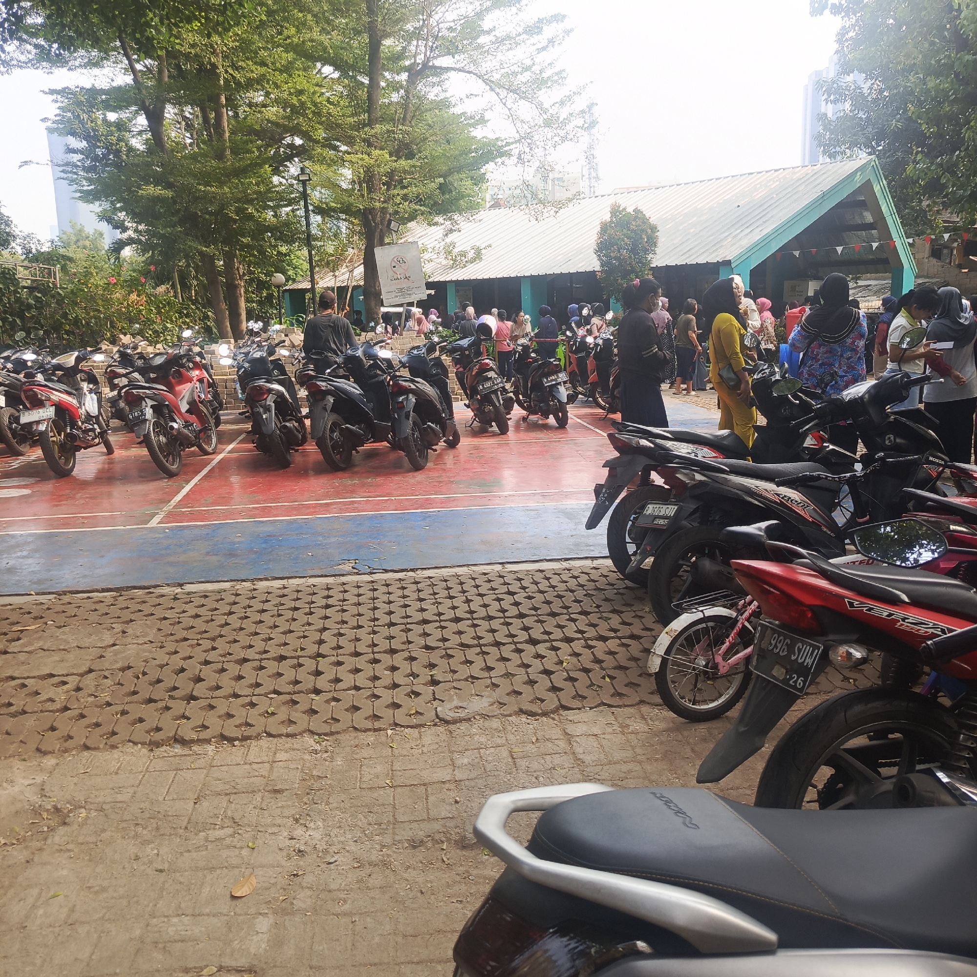 This image shows a group of people and a parking lot full of motorcycles in South Jakarta, Indonesia. The scene appears to be in an outdoor area with trees and a building in the background. The ground is a red and blue court with white lines, possibly a basketball court, surrounded by a paved area with interlocking bricks.
In the foreground, several motorcycles are parked. The closest motorcycle has a black seat and silver handlebars. Behind it, a red motorcycle with a license plate reading "B 996 SUW" is visible. Further back, more motorcycles of various colors, including red, black, and silver, are parked in rows.
A group of people, some wearing hijabs, are standing and walking around the motorcycles and the building. The building in the background has a light-colored roof and teal-colored walls. There is a sign visible on a pole near the building, with text that is difficult to read but appears to be a public notice. Tall trees provide shade over the area. In the far background, a tall modern building can be seen, suggesting an urban environment within South Jakarta. The overall atmosphere appears to be casual, with people gathering outdoors.