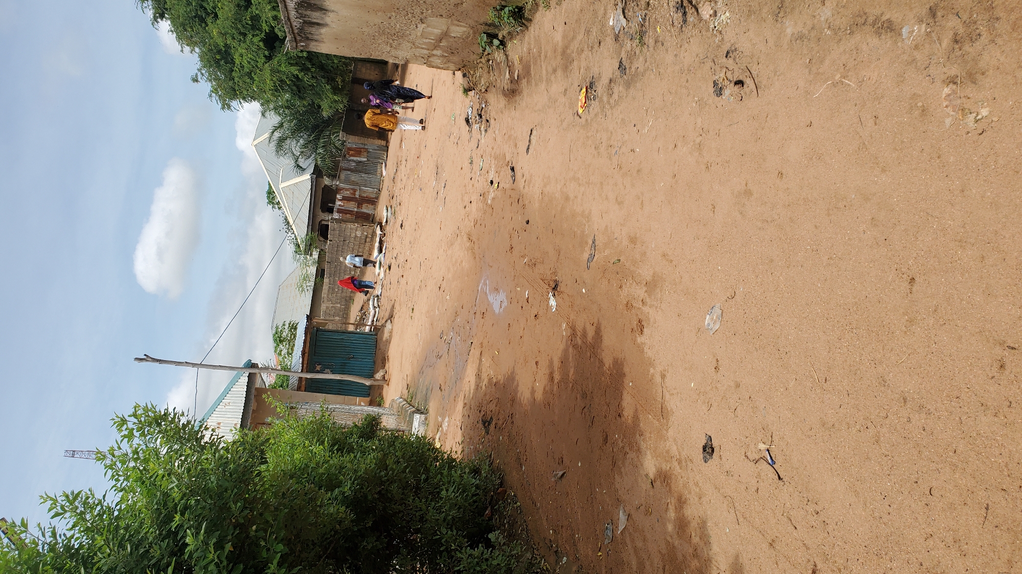 A wide shot captures a dusty, unpaved street in Jalingo, Nigeria, under a bright, cloudy sky. Several people are walking in the distance. On the left, a dense green tree overhangs the street. The street itself is littered with small pieces of debris. Buildings with corrugated metal roofs line the sides of the street, interspersed with walls constructed from concrete blocks and corrugated metal sheeting. One building features a teal-colored metal door. A thin wooden pole stands vertically near the teal door. In the background, a telecommunications tower is visible against the sky.
