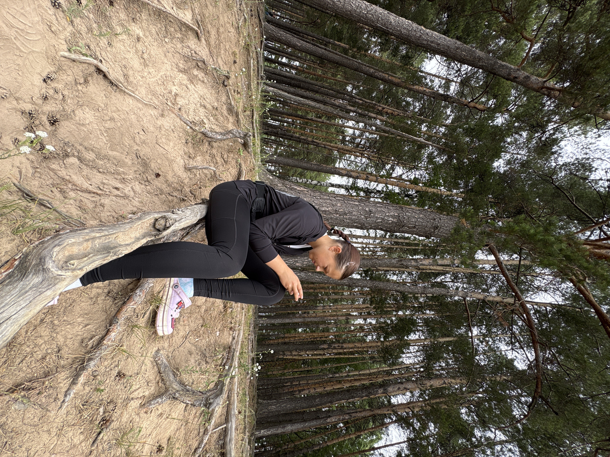 The image shows a young woman in black athletic wear and pink shoes, resting on a sandy incline with exposed tree roots. She is crouched, with her head bowed and hands clasped in her lap, appearing contemplative or tired. The background features a dense forest of tall pine trees, with their trunks running vertically due to the image's rotation. The ground is covered in sand, dry grass, scattered pinecones, and small white wildflowers. The lighting suggests it could be daytime, possibly overcast given the muted light. The overall scene is outdoors, in a natural, wooded environment. The woman's posture and the natural setting do not convey a specific event, but rather a moment of rest or pause. There is no visible text in the image.