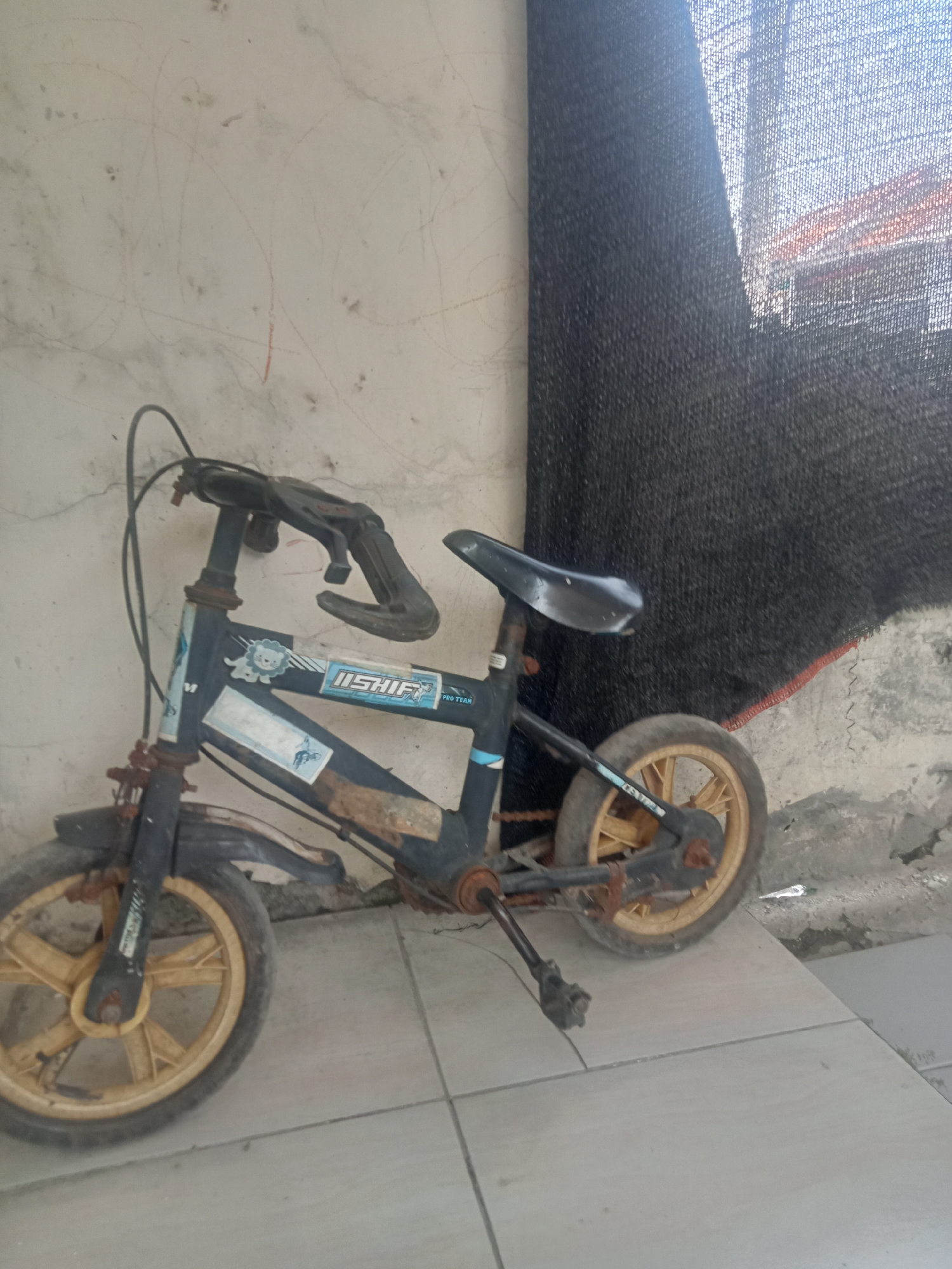 The image captures a weathered, dark blue children's bicycle with yellow plastic wheels, resting against a textured, light-colored wall. The bicycle exhibits signs of rust, particularly on the frame and spokes, and appears to be in a state of disuse. A black mesh fabric is draped behind the bicycle, partially obscuring the background, which reveals hints of buildings with red tile roofs, suggesting an outdoor setting. The ground is paved with large, square, light gray tiles. The overall impression is one of a forgotten object in an urban or semi-urban environment. While the location is stated as Kutosari, Indonesia, no specific landmarks are visible to confirm this. There are no people or animals present in the image.