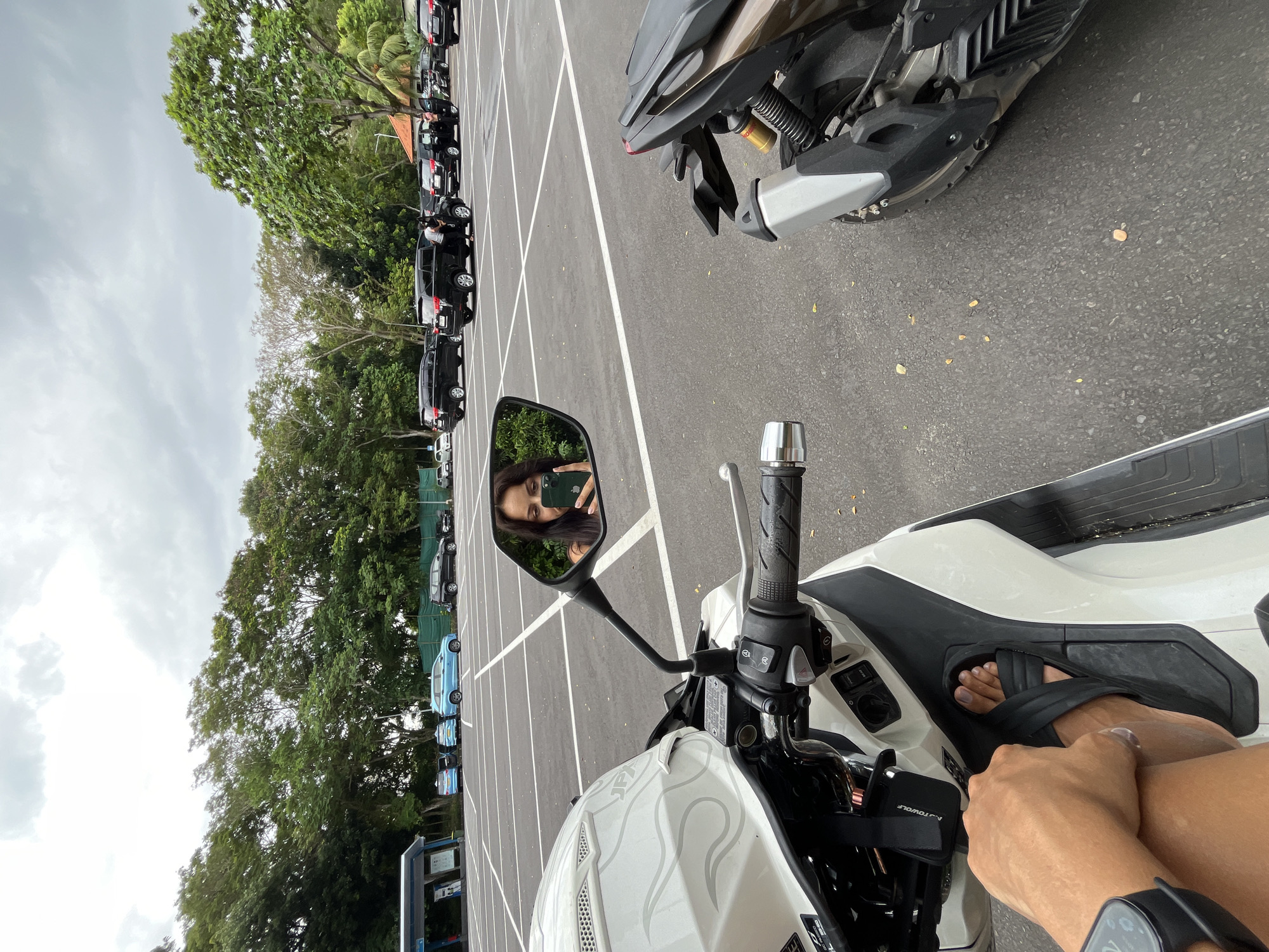 The image presents a first-person perspective from a person seated on a white scooter in a large outdoor parking lot. In the scooter's rearview mirror, the photographer, wearing sunglasses and holding a smartphone, is visible, capturing the scene. Their right arm, adorned with a smartwatch, and legs, featuring painted toenails and black sandals, are seen in the foreground, resting on the scooter's footboard. A white helmet with an embossed design is placed on the scooter's front. To the right, parts of another motorcycle, with dark and metallic components, are partially visible.
The spacious parking lot, made of asphalt with clear white painted lines, extends into the background, filled with numerous parked vehicles. The majority of these are black SUVs or vans, arranged in rows, with a few light blue cars visible further in the distance. A dense line of lush green trees, including some palm trees, borders the parking area under a bright, overcast sky, indicating daytime. The text "AUTOWOLF" is discernible on a component near the scooter's handlebars. The setting appears to be a public or tourist area in Kuta Selatan, Indonesia.