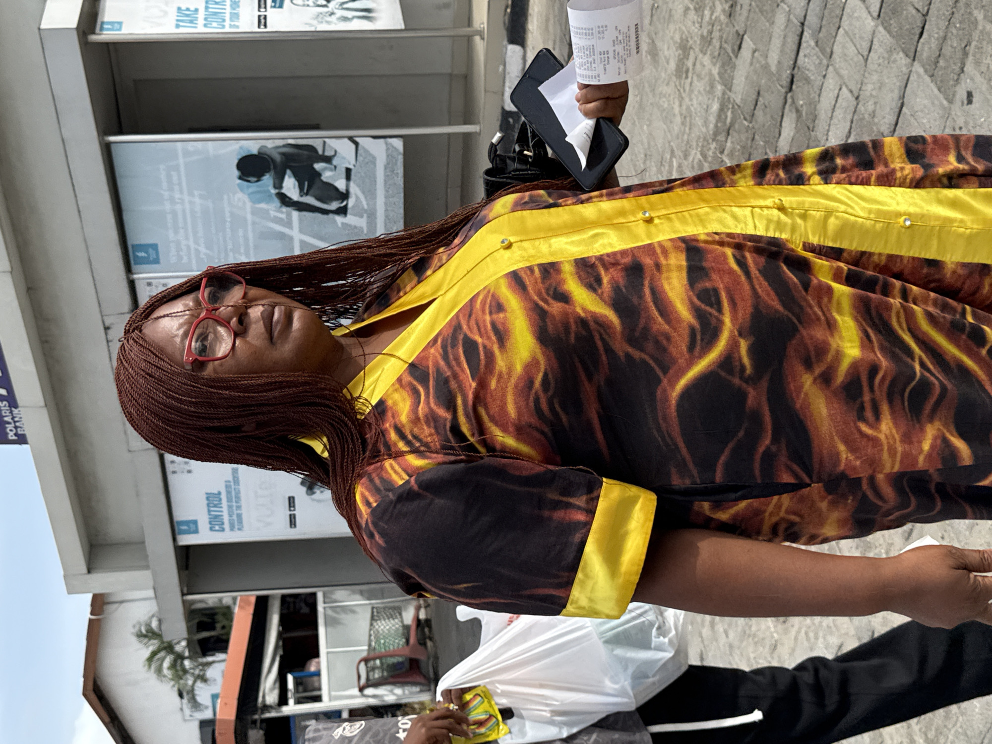 A woman with reddish-brown braided hair and red-framed glasses stands outdoors, facing forward with a neutral expression. She is wearing a long-sleeved dress or tunic featuring a black base with a yellow and orange flame-like pattern. A solid yellow band runs vertically down the front and horizontally along the sleeve cuffs, adorned with small, shiny studs.
In her right hand, she holds a black wallet or small purse, from which a rolled-up paper receipt protrudes. Her left hand is visible further down, holding a white plastic bag that contains a colorful item, possibly yellow and green.
The background shows a light-colored building, likely a commercial establishment, with several posters and signs. Text visible on these signs includes "TAKE CONTROL," "CONTROL," "VOLT," and "POLARIS BANK." One poster depicts a person in a dynamic pose. The ground is paved with light-colored tiles. A small palm-like plant is visible in the middle ground. The clear blue sky and bright lighting suggest it is daytime. Part of another person's leg and foot, wearing black pants, can be seen at the bottom right of the frame.