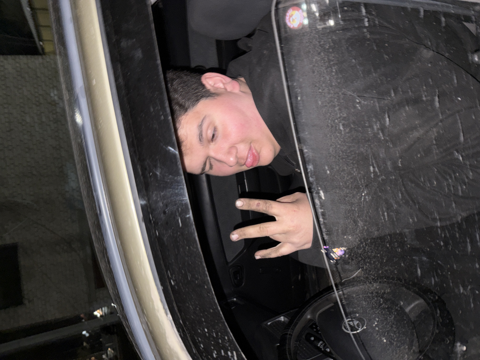This image, captured in Chaikovsky, Russia, shows a young male leaning out of the driver's side window of a car, likely at night or during the evening. The individual has dark hair and light skin with prominent rosy cheeks, and is wearing a dark, possibly black, hooded jacket or sweatshirt. He is looking towards the camera with a relaxed and slightly playful expression, his tongue gently visible. His right hand is extended, making a gesture with the index, middle, and ring fingers raised.
The interior of the car is dark, with a portion of the steering wheel visible at the bottom right. The car window appears streaked with dirt or moisture, possibly indicating rain or general grime. A small, circular sticker featuring a cartoon-like face or character is affixed to the upper right corner of the window. The background outside the car is indistinct and dark, with blurry reflections suggesting distant urban structures or lights. The photograph is taken from outside the vehicle, looking in and slightly downwards, with parts of the car's frame visible on the left and right edges. No discernible text is visible within the image.