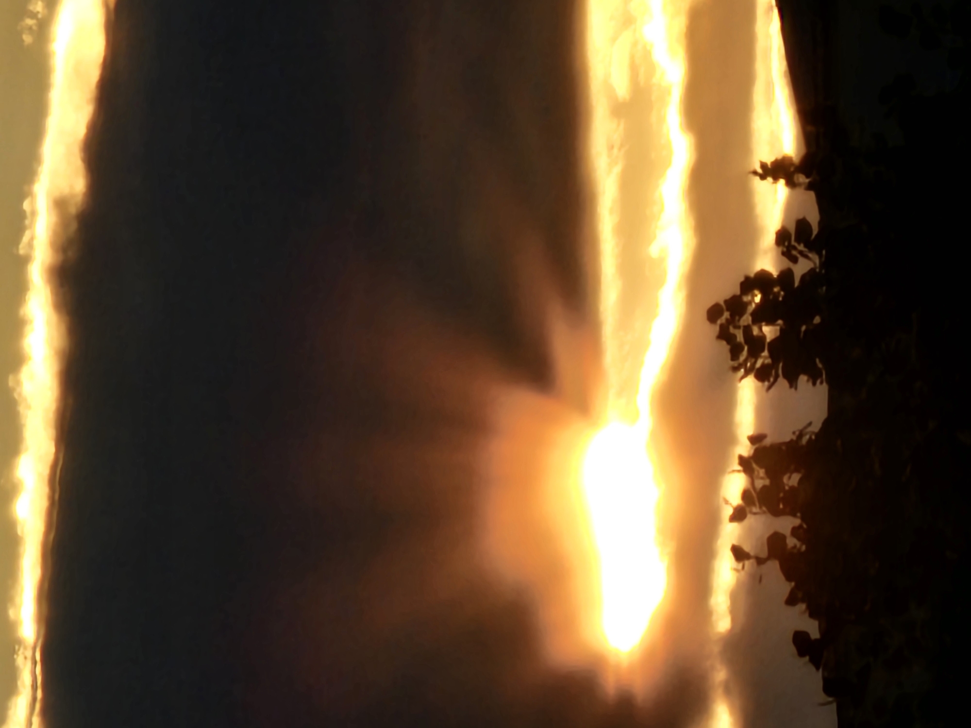 The image is a vertical, dimly lit photograph taken at dusk or dawn, featuring a dramatic sky with the sun partially obscured by dark clouds. The sun's rays emanate upwards, creating a luminous effect. In the foreground, silhouetted trees and foliage create a dark, indistinct border against the brighter sky. The scene appears to be an outdoor landscape, possibly a park or a residential area in Khimki, Russia, given the context. There are no visible people or specific identifiable landmarks. The overall impression is one of natural beauty and atmospheric lighting.