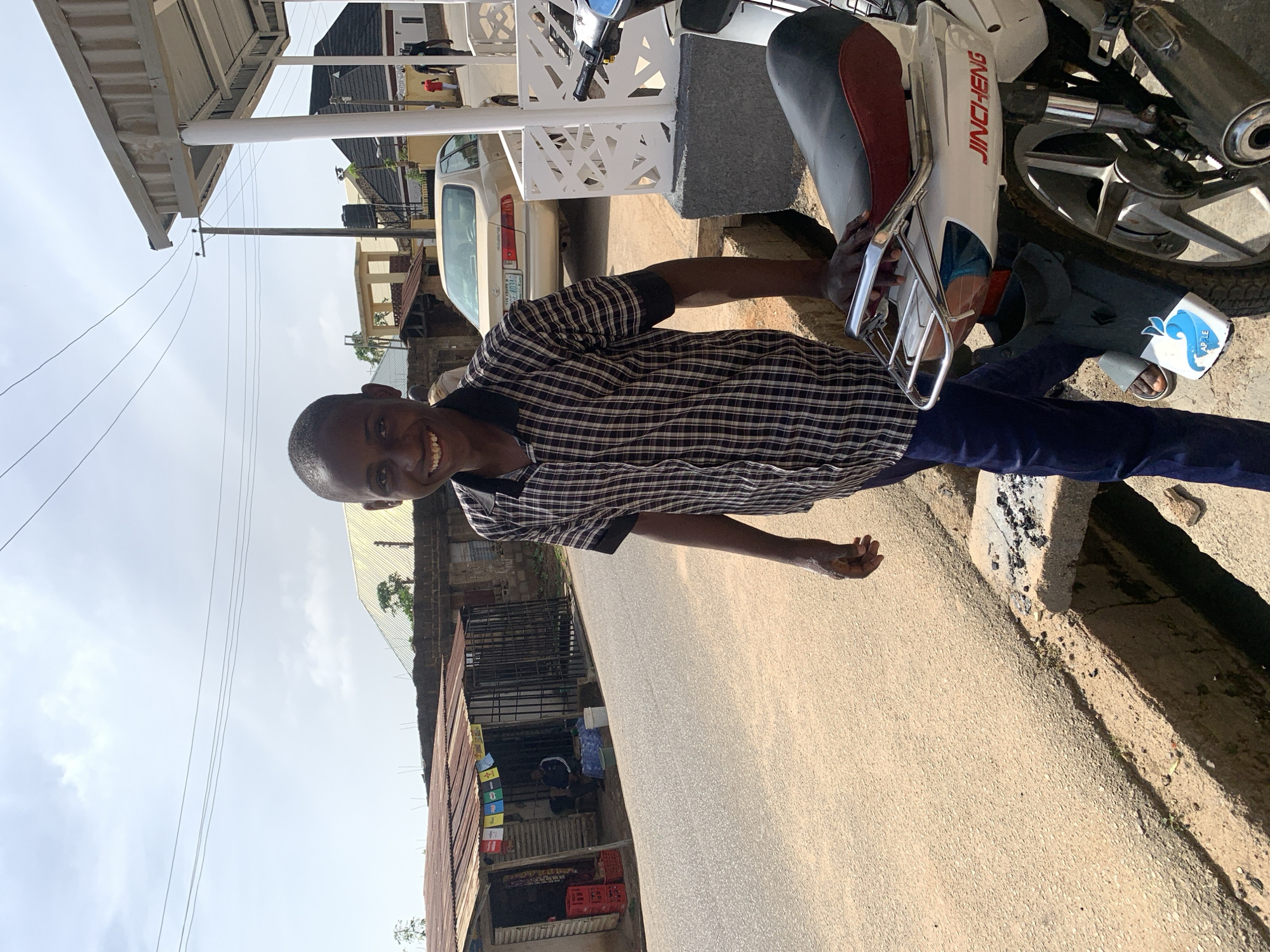 A young Black male stands in front of a white motorcycle on a sunny day in Jos, Nigeria. He is smiling broadly at the camera. He is wearing a checkered shirt and dark blue pants. The motorcycle has red accents on the seat and "JINCHENG" written in red on its side. In the background, there are buildings with corrugated metal roofs, a street, and other vehicles. The scene appears to be in a commercial area.