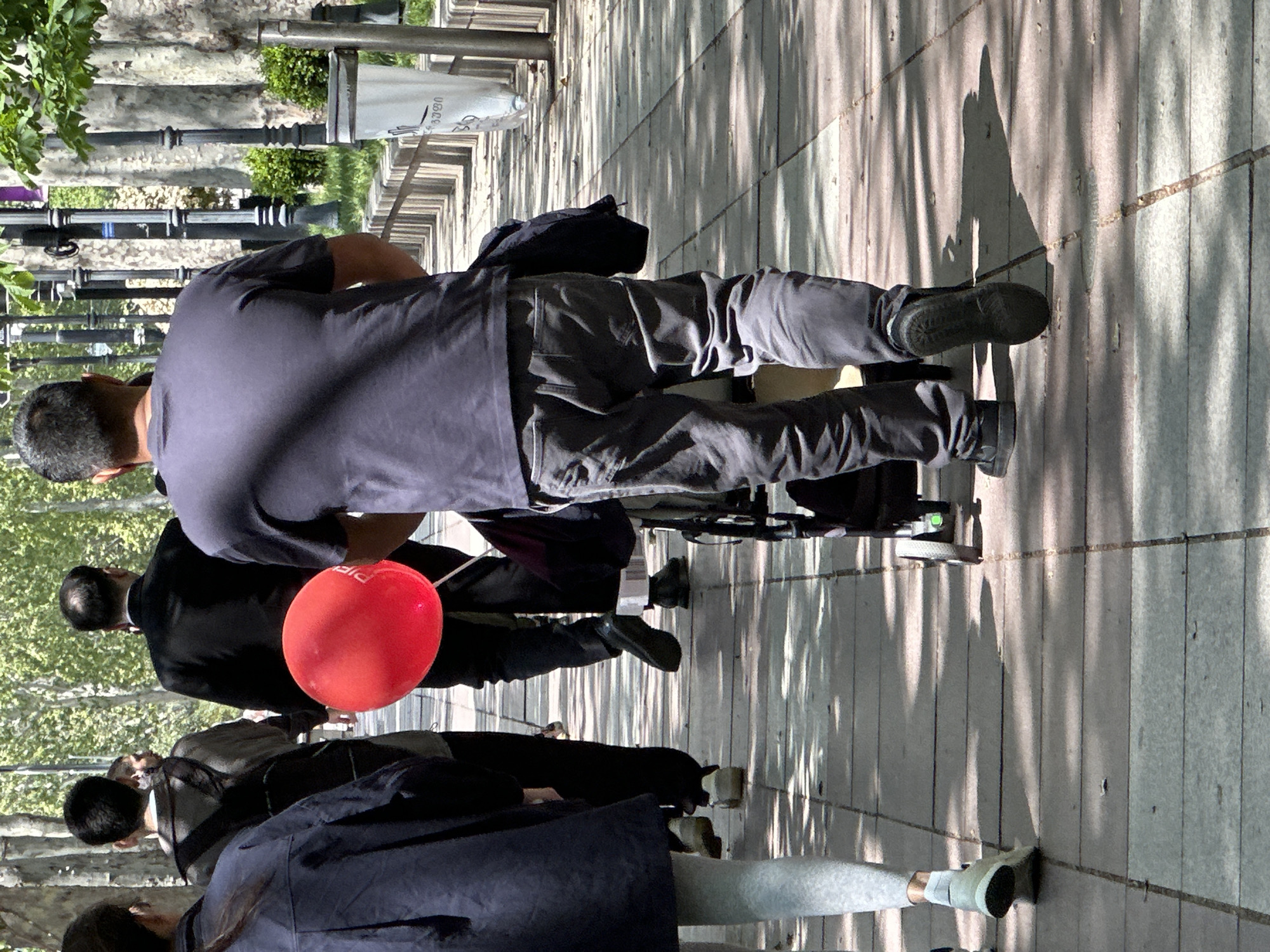 The image captures a street scene in Tbilisi, Georgia, with people walking along a sidewalk. The sidewalk appears to be constructed of concrete slabs with long grooves. A group of pedestrians are present, including individuals wearing casual clothing such as jeans and dark shirts, with one person holding a red balloon. In the background, there are trees, lamp posts, and greenery, suggesting an urban park or a tree-lined street. The overall atmosphere suggests a sunny day with shadows cast by the surrounding trees.