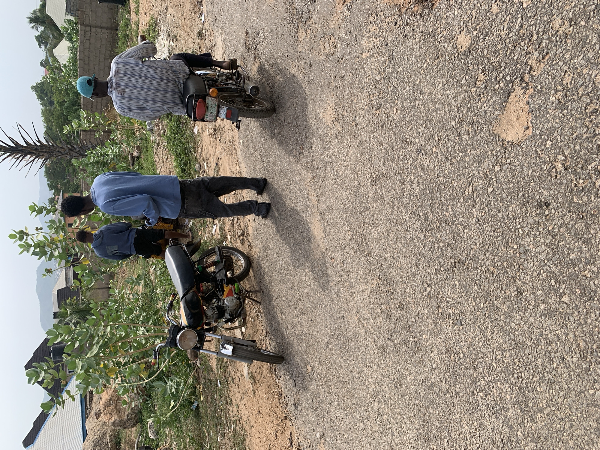 The image shows two men standing beside two motorcycles on a paved road in a semi-urban area, likely in Lokoja, Nigeria. One man is wearing a striped shirt and a blue cap while sitting on a motorcycle. The other man stands beside his motorcycle, wearing a blue shirt and dark pants. The road shows signs of wear and patching. The background includes trees and vegetation on one side and buildings in the distance. The scene appears to be in daylight.