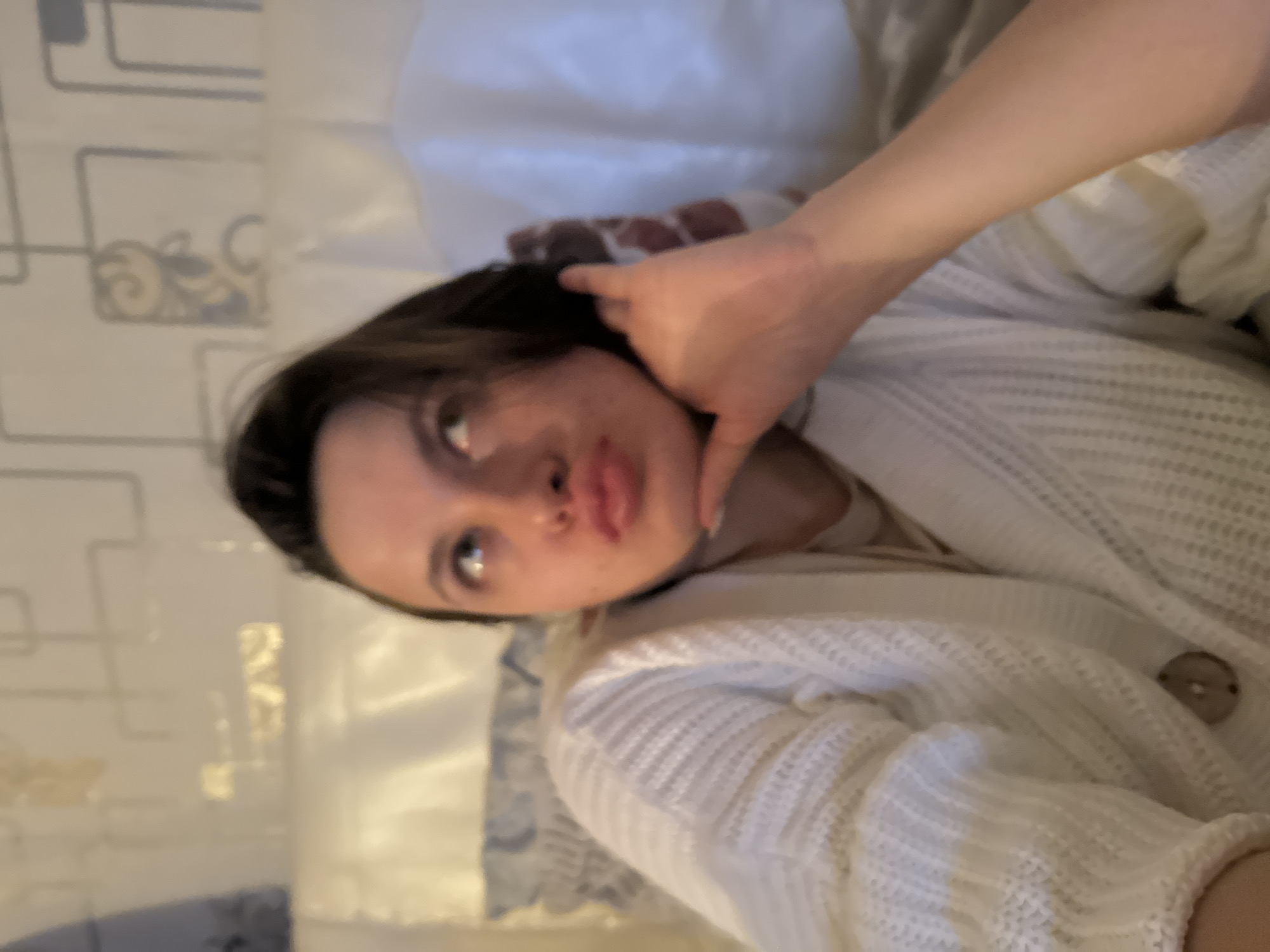 A medium shot shows a fair-skinned woman with dark hair, lying in bed, wearing a white sweater. Her left hand is near her face. The room features white bedding and patterned wallpaper. The location is unknown.