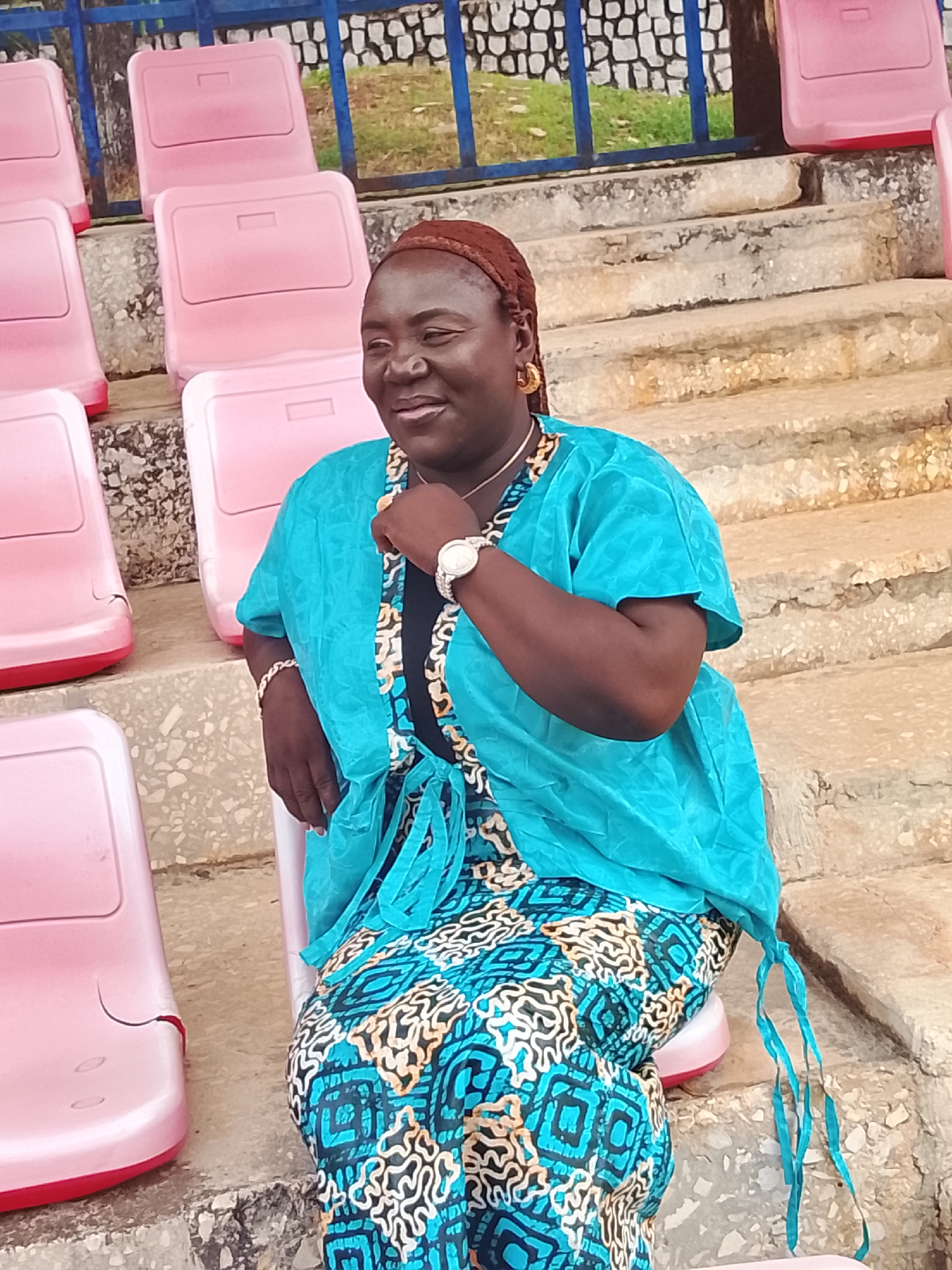 A middle-aged African woman wearing a blue and patterned dress and a blue blouse is seated on a pink seat in a stadium-like setting. She has dark skin, a brown headscarf, and gold jewelry. She is looking to her left and resting her chin on her hand. The background features rows of pink seats, concrete steps, and a blue metal railing. The location is Zawan, Nigeria.