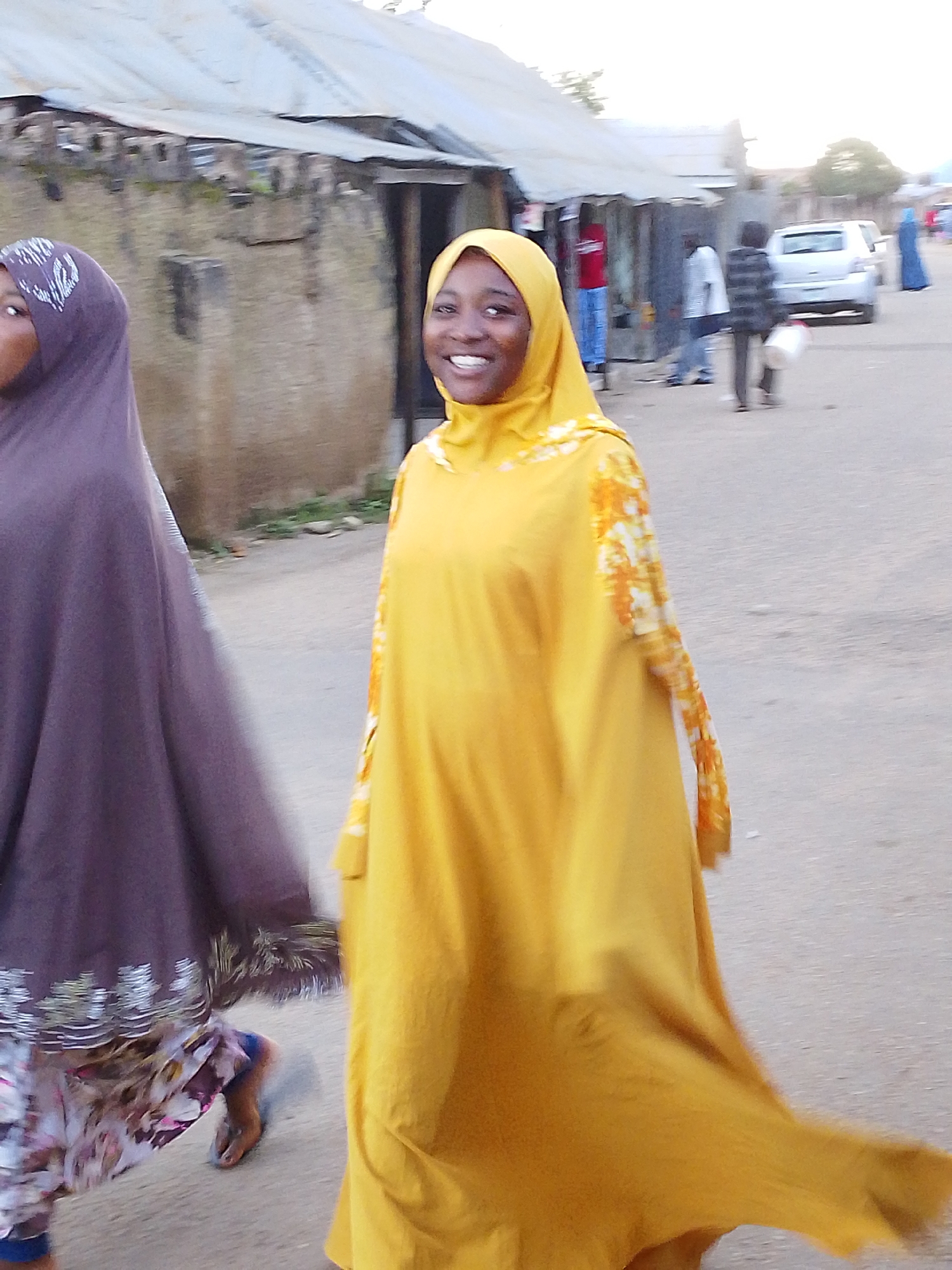 In Jos, Nigeria, two women walk on a dusty street. The woman on the left wears a dark purple hijab and a patterned skirt. The woman on the right wears a bright yellow hijab and a matching yellow dress with orange floral accents on the sleeves. Her face is visible, and she is smiling. In the background, there are buildings with corrugated metal roofs, and other people and a car are visible further down the street. The overall scene depicts a casual street view in a Nigerian city.