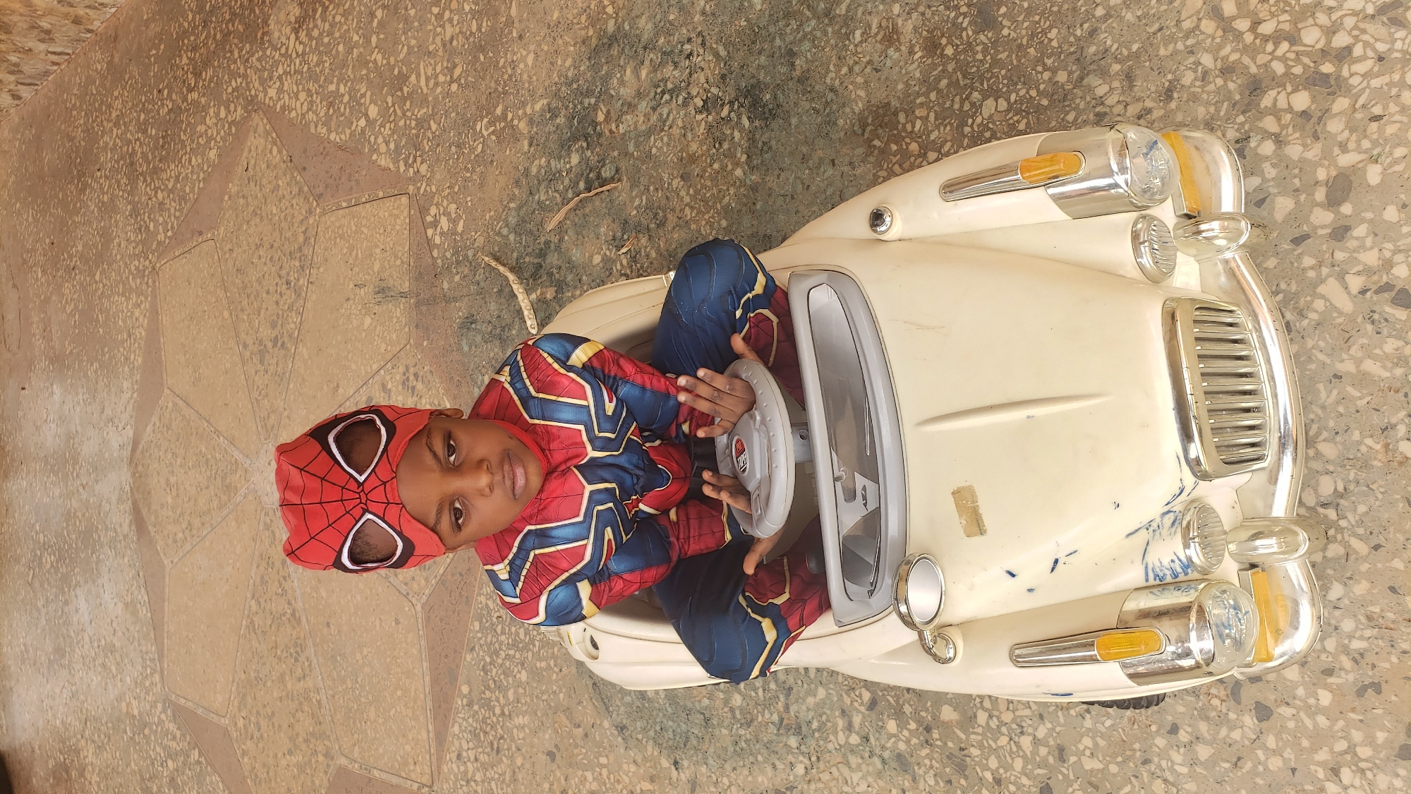 A young child wearing a Spider-Man costume is sitting in a white toy car, gripping the steering wheel. The scene appears to be outdoors, possibly on a tiled or paved surface. The toy car is a vintage model with chrome details. The location is possibly in Sabon Tasha, Nigeria.