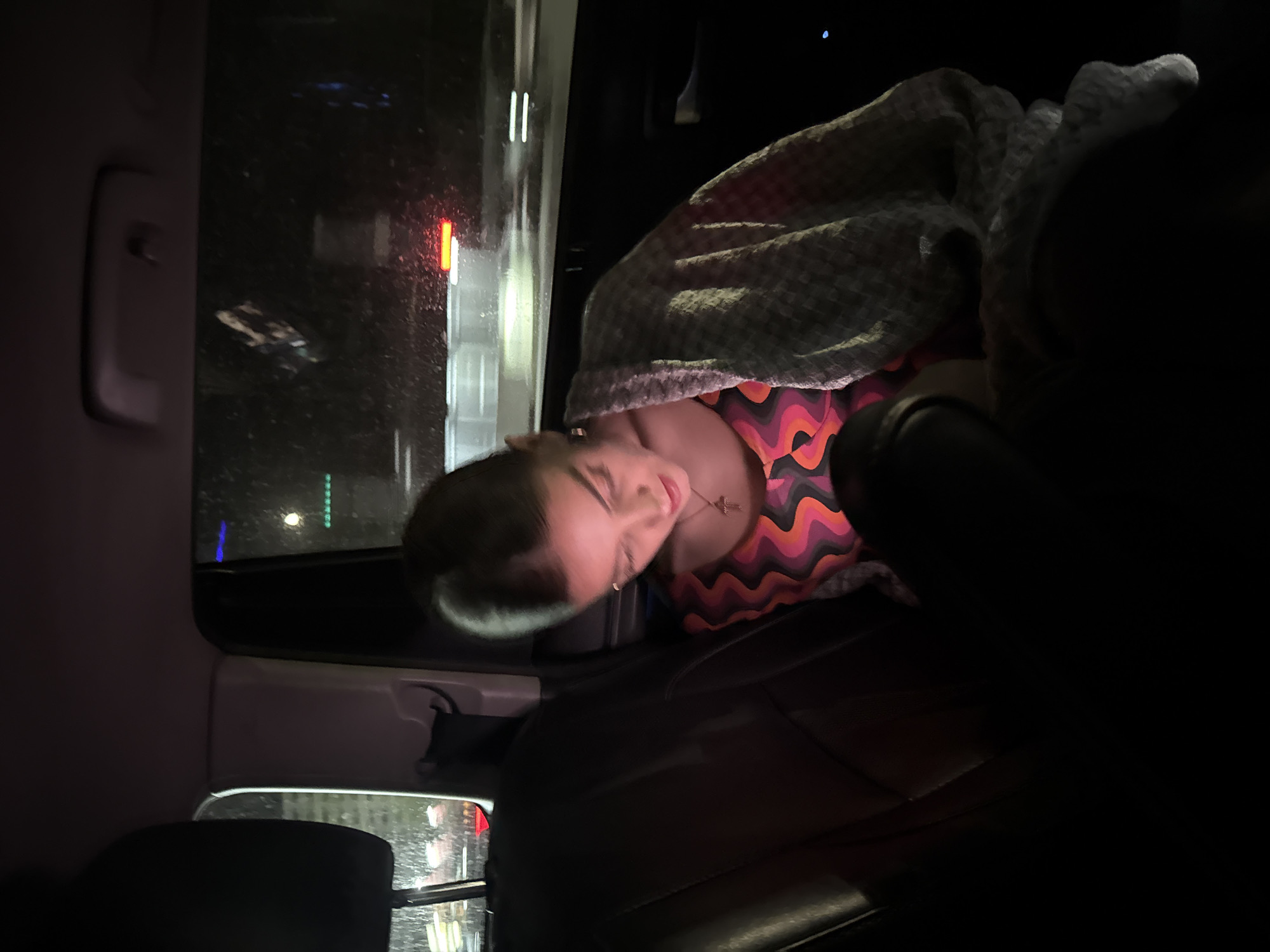 A person is sleeping in the back seat of a vehicle at night. The individual is covered with a checkered blanket and wearing a colorful, wavy-patterned top. The vehicle appears to be in a city setting, with lights visible outside the window. The specific location is unknown.