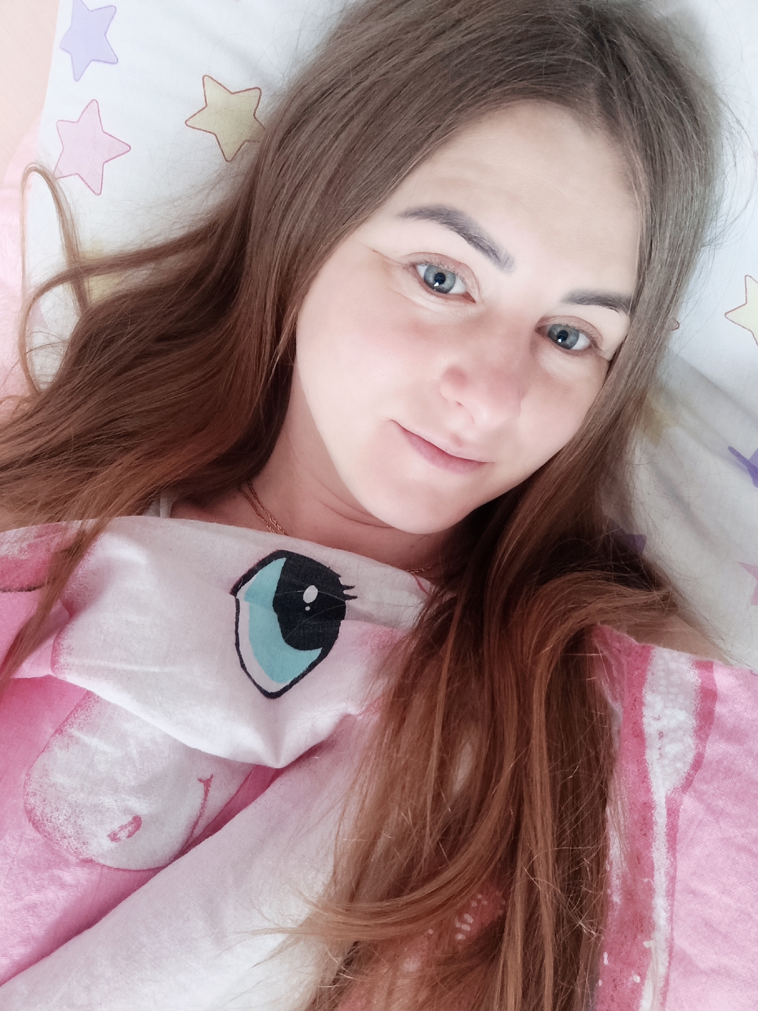 A young woman is depicted in a close-up, eye-level shot, appearing to lie in a bed. She has long, wavy brown hair, which is fanned out around her head and shoulders. Her eyes are light blue, and she looks directly towards the camera with a soft, gentle smile. Her skin is fair, and she has well-defined eyebrows.
She is partially covered by a pink and white blanket or duvet, which features a large cartoon eye design on the visible portion, reminiscent of characters like My Little Pony. The eye is predominantly black and light blue with a white highlight, outlined in black. A thin, gold-colored chain necklace is visible around her neck, resting on her chest.
Underneath her head is a white pillow adorned with pastel-colored star patterns in purple, pink, and yellow. The overall setting suggests an indoor environment, likely a bedroom, given the presence of a bed, pillow, and blanket. The lighting is soft and even, not indicating a specific time of day. The woman appears relaxed and at ease. There is no visible text in the image.