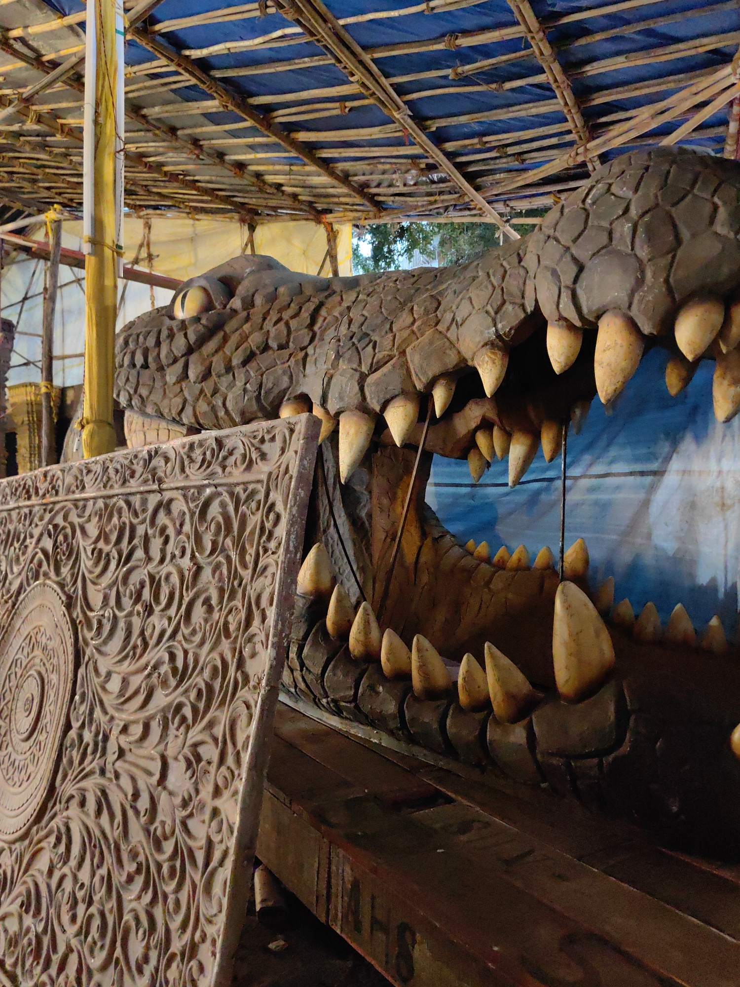 The image depicts a large, artificial crocodile head with its mouth wide open, revealing numerous sharp, yellowish teeth. The crocodile appears to be part of a temporary structure, possibly a decorative element for an event or festival. The background shows a rudimentary canopy made of bamboo poles and blue tarpaulin, suggesting an outdoor setting. To the left, a carved wooden panel with intricate floral and circular motifs is visible, leaning against the crocodile. The scene appears to be an outdoor setting in Mumbai, India, possibly a street fair or a Durga Puja pandal setup. The lighting is artificial, giving the scene a somewhat theatrical ambiance. There are no people visible in the frame.