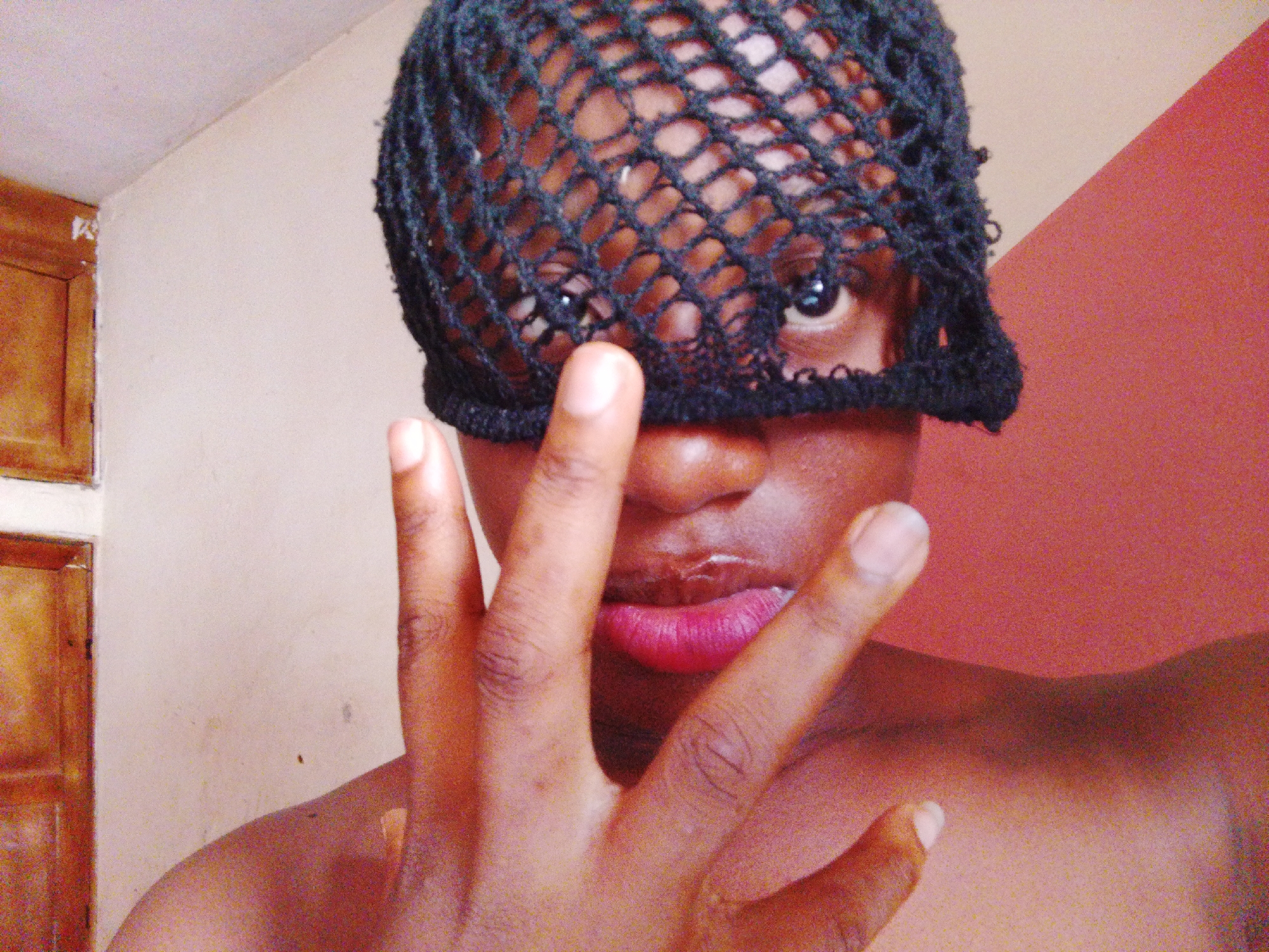 A person with dark skin is shown from the chest up, with a black net covering the forehead and eyes. A hand with long fingers is held up in front of the person's mouth and nose. The background is a wall with two different colors: tan and reddish-brown. A brown wooden door is slightly visible on the left side of the frame. The location is possibly in the city of Umuahia, Nigeria.