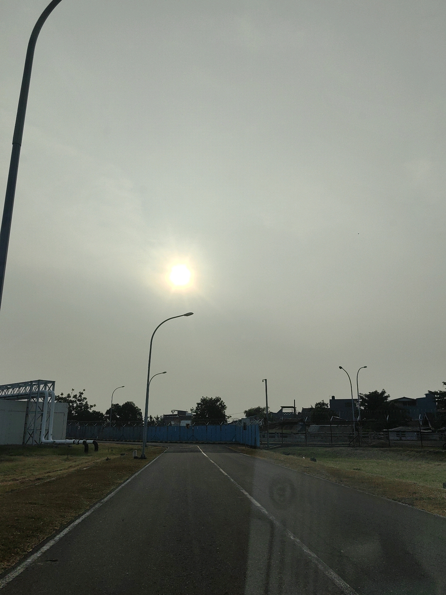 A paved road leads towards a blue gate with a fence and buildings behind it, in what appears to be an industrial area in Betro, Indonesia. The sky is overcast with the sun shining brightly through the clouds, creating a hazy atmosphere. Streetlights are visible along the road, and to the left, there is a building with pipes and a metal structure. Green grass lines the sides of the road. The image is taken from a low angle, possibly from inside a vehicle.