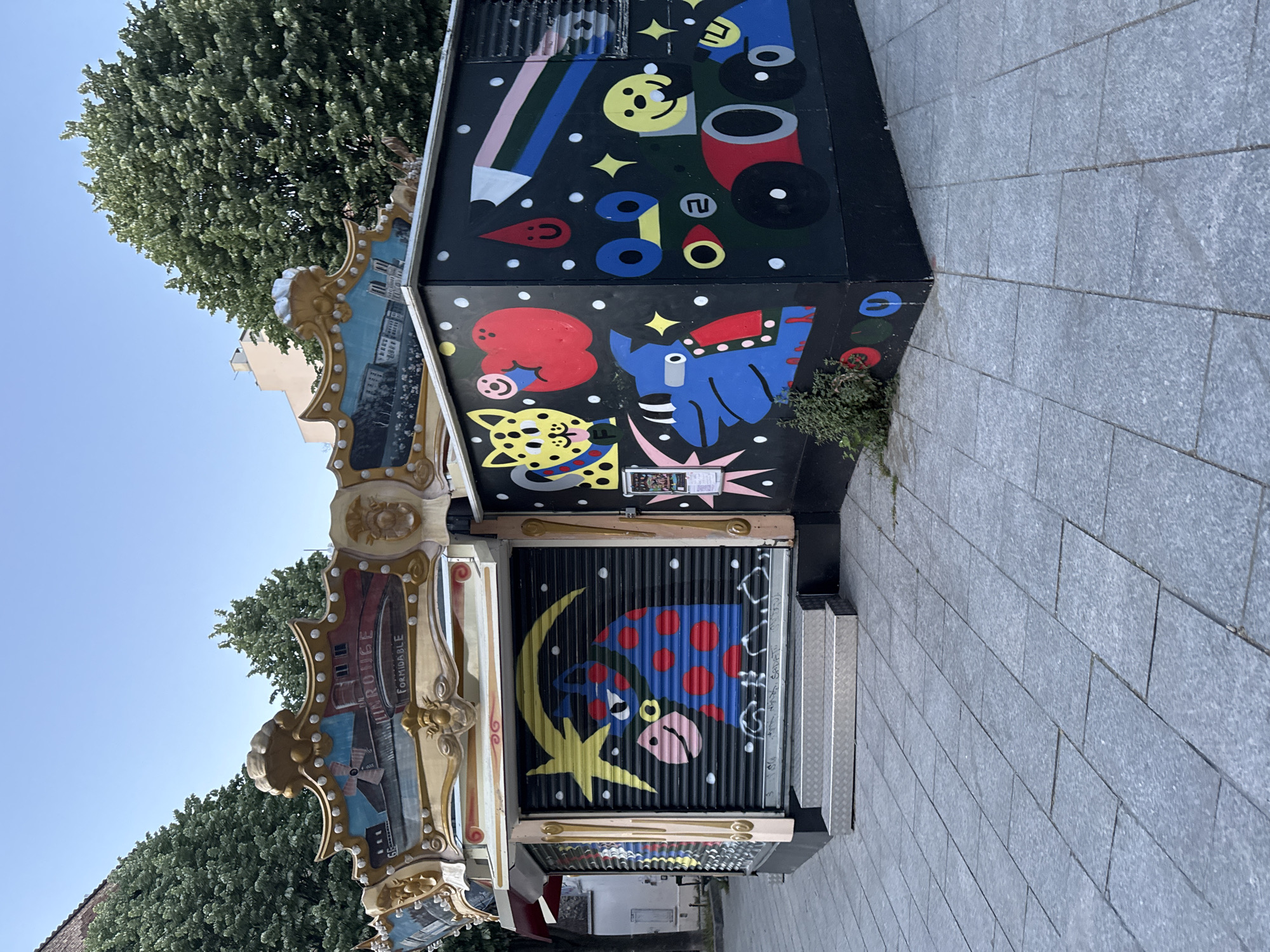 This outdoor image, captured in Paris, France, showcases a vibrantly decorated kiosk or stall under a clear blue sky. The structure is composed of two distinct parts: an ornate, vintage-style facade on the left and a modern, black-painted section on the right.
The left side of the kiosk features intricate, gilded carvings and decorative panels, reminiscent of a carousel or an old fairground attraction. One panel depicts a city scene in muted tones, and visible text includes "ENROUGE" above "FORMIDABLE".
The larger right section of the kiosk is painted black and adorned with bold, colorful, cartoon-like murals. The uppermost part displays abstract shapes, a large multi-colored pencil, a yellow smiley face, red and blue geometric forms, and scattered white stars, creating a whimsical effect. Below this, the mural continues with a yellow, spotted leopard-like creature, a red cloud-like shape with a smiley, and a blue stylized animal head, possibly a horse or elephant, also punctuated with stars and colorful details. The bottom part of the kiosk, which is a corrugated metal shutter, features a large blue creature with red polka dots and a pink face, positioned next to a yellow crescent moon and a star.
Lush green trees line the background against the bright sky, and the ground in the foreground is paved with light grey stone tiles. Metallic steps are visible on the far right, leading up to the kiosk. The scene is well-lit, indicating daytime with clear weather. No people or activities are visible, suggesting the stall is closed. The overall impression is one of playful urban art and historical charm.