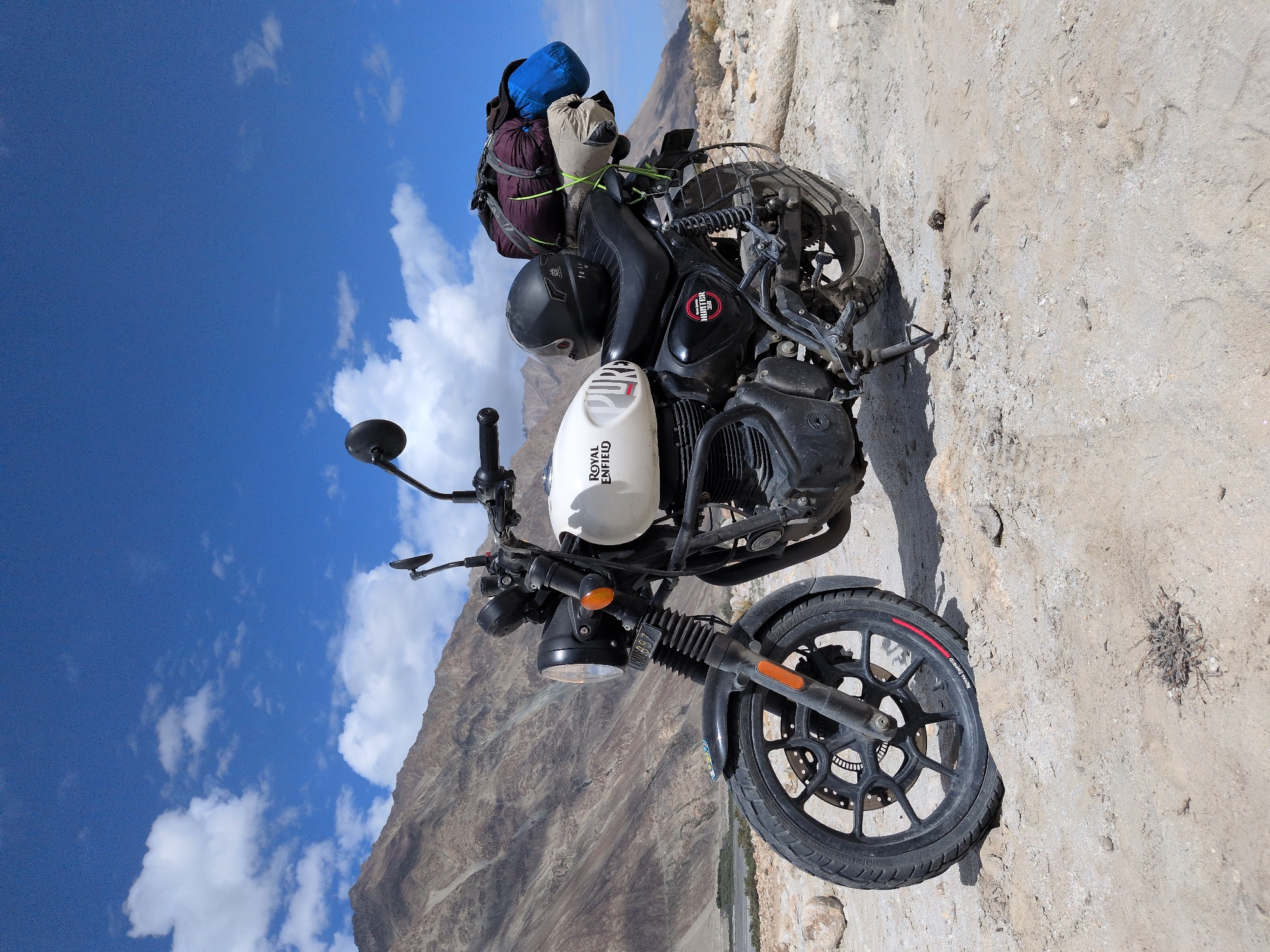 A Royal Enfield Hunter 350 motorcycle is parked on a rocky, sandy terrain. The motorcycle is white with "ROYAL ENFIELD" and "HUNTER" logos visible. It is equipped with saddlebags packed with camping gear, including a helmet. The background features arid, mountainous landscape with a clear blue sky dotted with white clouds. The location is identified as Sasoma, India.