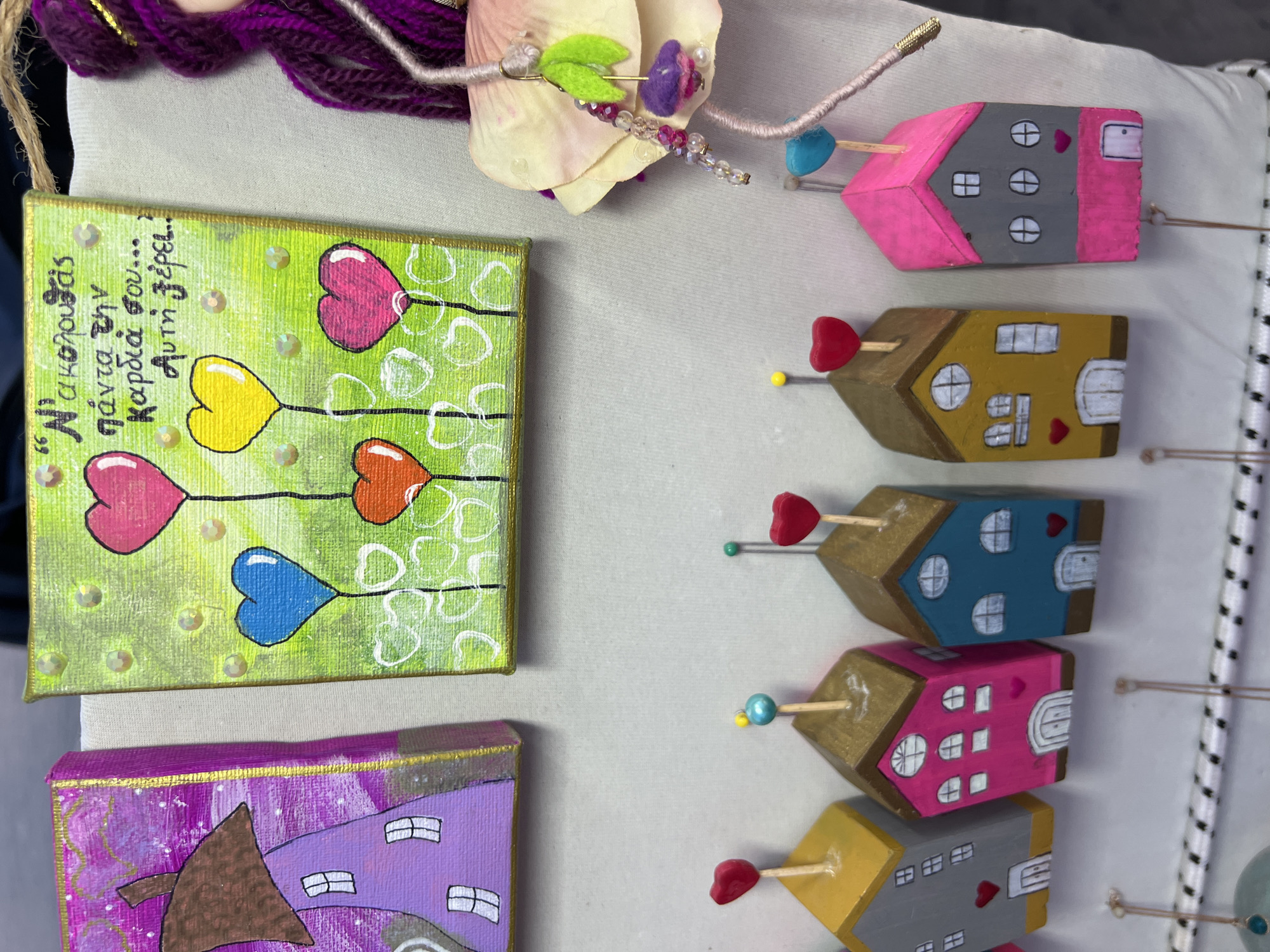 The image shows a close-up of various handmade crafts displayed on a white surface, likely a market stall. In the top half, there are two small square canvases with paintings. The left canvas features a purple house with a brown roof and white windows. The right canvas has a bright green background with several colorful heart-shaped balloons rising from a base decorated with small white hearts. Greek text is written in black script on this canvas. To the right of the canvases, a section of a doll with purple yarn hair and a pink and yellow fabric flower is visible.
The bottom half of the image showcases a row of six small, wooden house-shaped ornaments, each with a different color and a small decorative element on top, such as a red heart or a blue bead. These ornaments are hanging on a patterned cord. The overall setting suggests an outdoor or indoor market display of artisanal goods. The specific location in Eretria Municipal Unit, Greece, is not ascertainable from the visual information provided in the image.