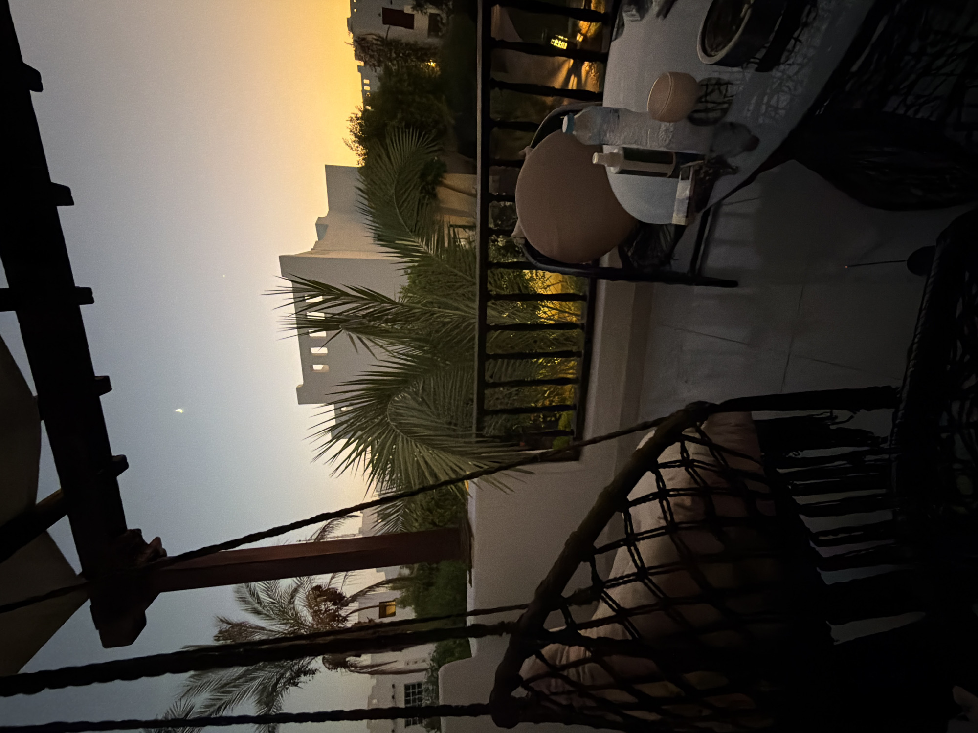 The image depicts a balcony at dusk in Sharm El Sheikh, Egypt, overlooking a resort complex. A dark hammock chair with cushions is in the foreground, hanging from a wooden beam. To the right, a dark-colored table is set with a bottle of water, a pump bottle, and a small bowl. Beyond the balcony, palm trees and white resort buildings are visible against a gradient sky of pale blue to orange. A crescent moon is visible in the upper left quadrant of the sky. The lighting suggests it is either sunrise or sunset.