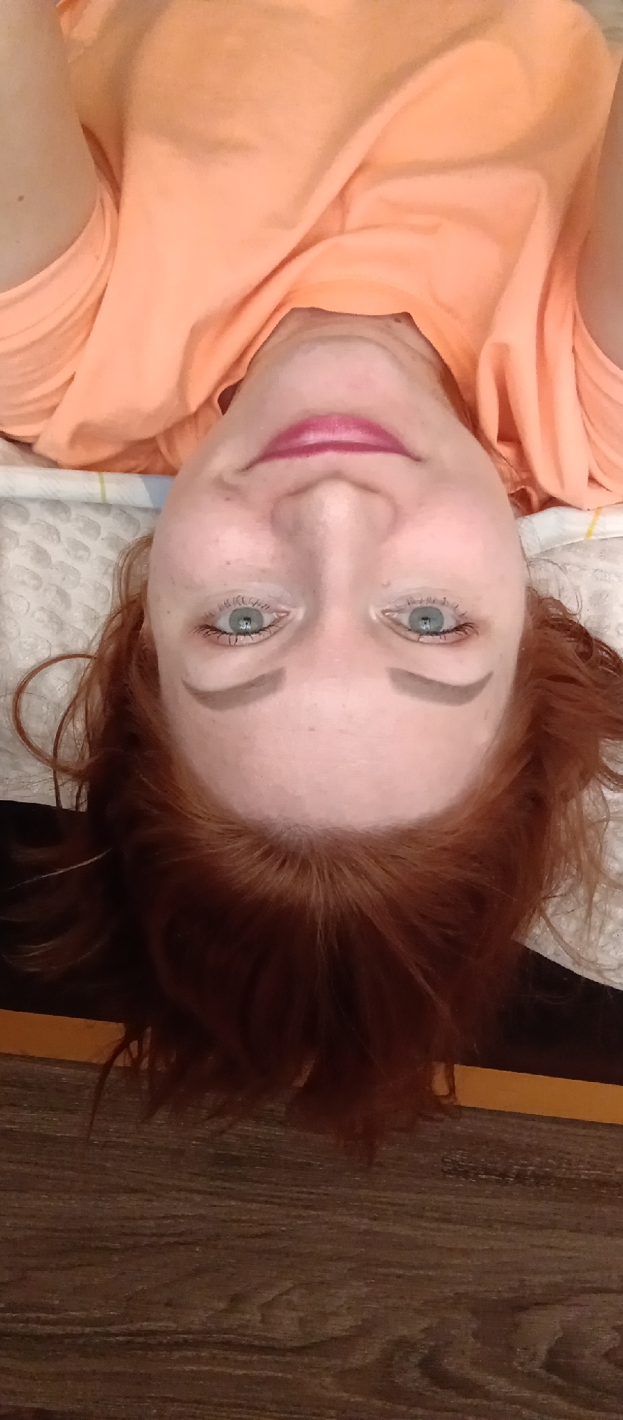 An upside-down selfie of a woman with red hair, green eyes, and pink lipstick, wearing an orange shirt and lying on a patterned surface resembling a pillow or blanket. The lower portion of the image shows a dark, wood-like surface. The location and the city are unknown.