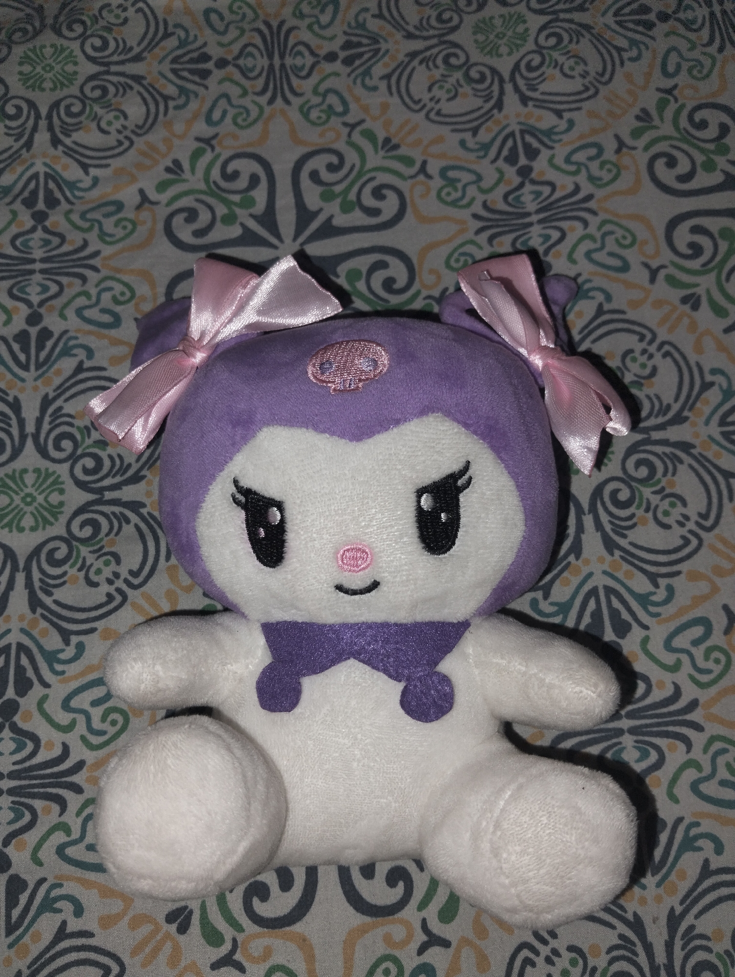 A plush toy, resembling the character Kuromi, is centered and depicted in a sitting position on a patterned fabric surface. The toy features a white plush body and face, contrasted with a purple plush hood covering the head, which includes two pointed ears, each adorned with a pink satin bow. A small, pink embroidered skull emblem is visible on the center of the purple forehead. The toy's face displays black embroidered almond-shaped eyes with small white highlights, a pink embroidered oval nose, and a simple black embroidered curved mouth. A purple plush bow-like element is attached to the toy's chest. Soft shadows are cast to the toy's lower right, indicating an indoor lighting source. The background fabric exhibits a repeating ornamental pattern of dark grey, green, and light orange curvilinear motifs against a light beige ground. No discernible features within the image indicate a specific location in Dhaka, Bangladesh.