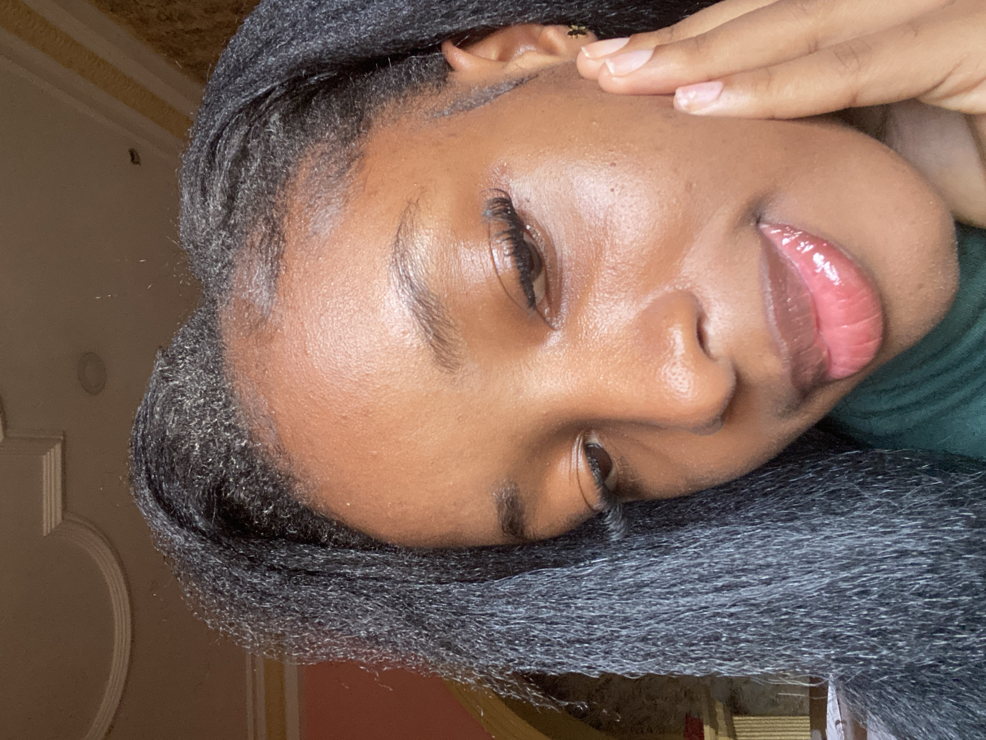 A close-up, slightly low-angle portrait features a young individual with dark brown skin. The subject's long, dark hair, styled in braids with visible texture, frames the face, parting on the left. The right eye is partially closed and looking downwards, accentuated by long, dark eyelashes. Eyebrows are dark and well-defined. The nose is visible on the right side of the face, and the full lips are glossy with a pink-red hue. The skin exhibits a visible sheen on the forehead, cheeks, and lips. The subject's right hand is raised, with fingers gently touching the right cheek. A small, dark earring, possibly butterfly-shaped, is visible on the right earlobe. A dark green garment is discernible around the neck area. The background indicates an indoor setting with light-colored walls and ceiling featuring decorative architectural molding. The image does not contain discernible visual information to confirm the specific location as Jos, Nigeria.