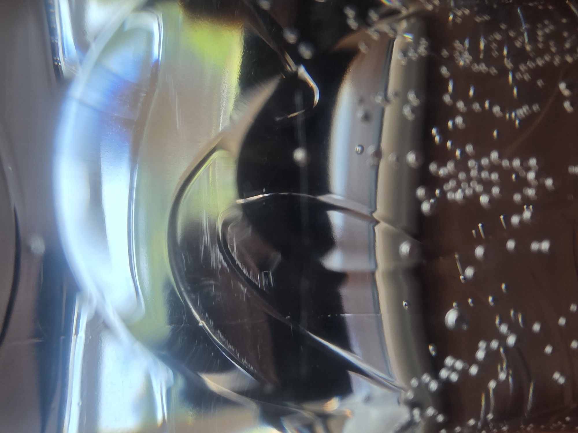 The image displays a close-up view of a clear plastic bottle filled with a clear liquid, likely water or a carbonated beverage, as evidenced by the numerous small bubbles present. The bottle has a ridged texture, creating a series of raised and recessed areas that distort the light and refract colors, producing a prismatic effect in some sections. The background appears to be a dark, possibly wooden surface, with some areas of green visible, suggesting an outdoor or brightly lit environment. There are visible scratches on the surface of the bottle, indicating wear or damage. The image does not provide any information that would allow for its location to be determined within the city of Ryazan, Russia, or any other specific geographic location.