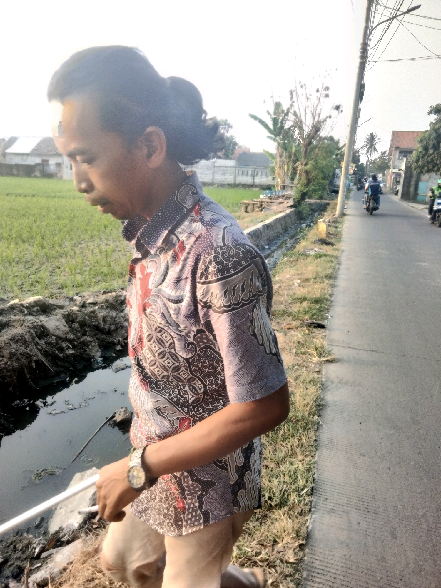 A man with his hair tied in a bun stands next to a muddy ditch. He is wearing a patterned shirt and light-colored pants. He holds a white pipe in his left hand and wears a watch on his right wrist. In the background, there is a field of green crops, houses, and a road with several motorcycles. The scene appears to be in a rural or suburban area, possibly the city of Suka Sari, Indonesia. The lighting suggests it is daytime, possibly late afternoon given the shadows.